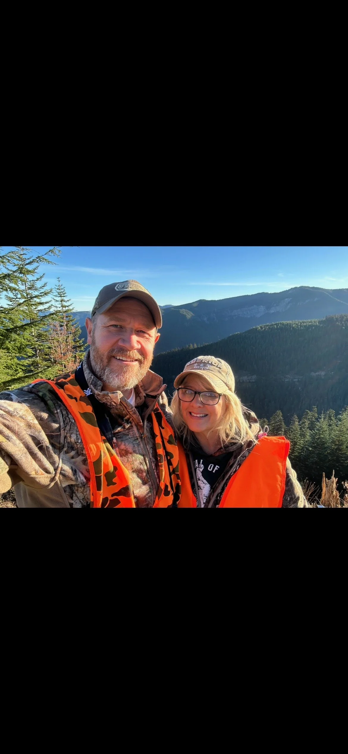 A man and woman taking a selfie outdoors in a mountainous forest area, both wearing hunting or outdoor gear, with green pine trees and mountain ranges in the background on a sunny day.