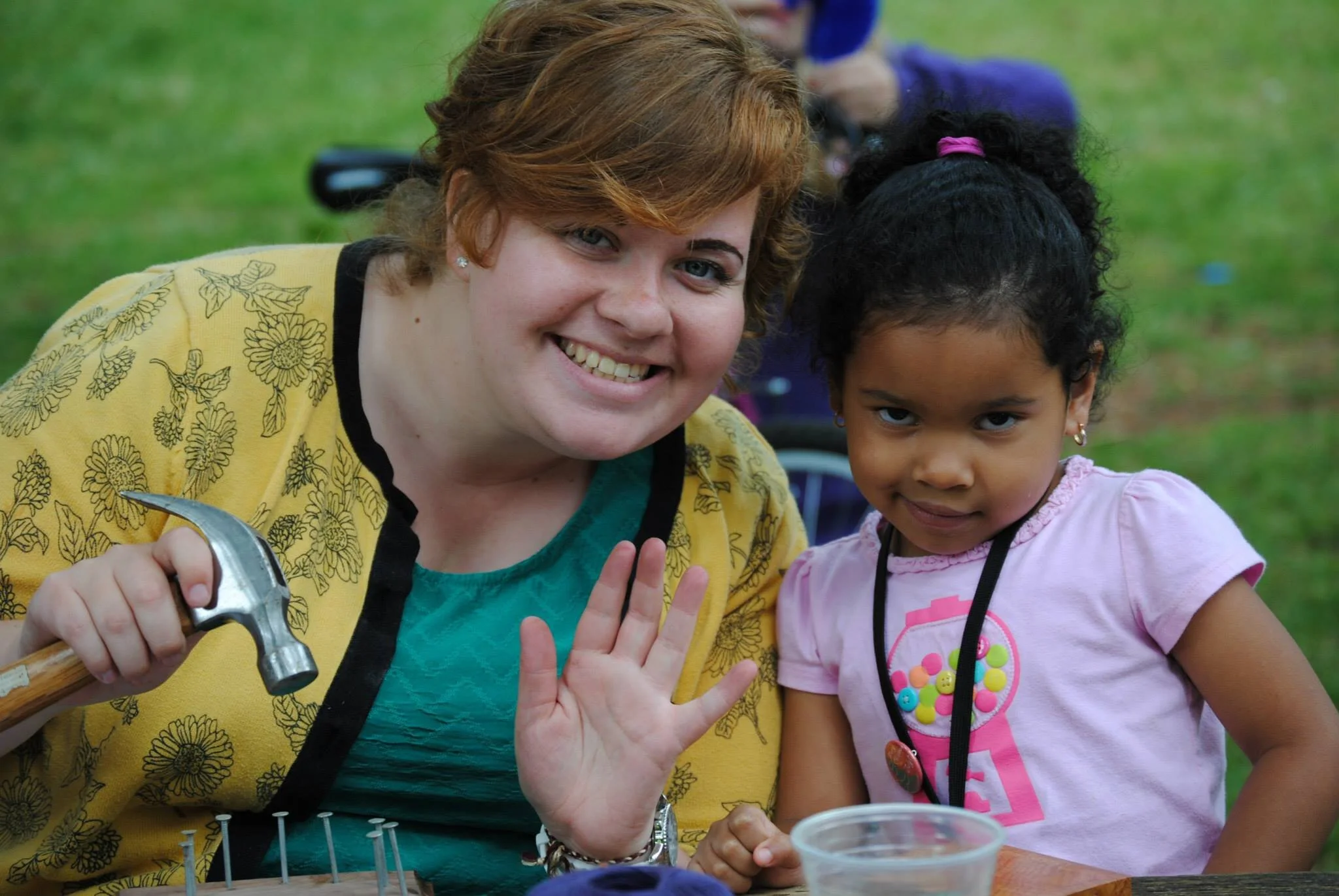 A young woman with short red hair and a girl with curly black hair are smiling at the camera. The woman is holding a hammer and has her hand raised as if waving. The girl is wearing a pink shirt with colorful buttons. They are outdoors on a grassy area, participating in a craft activity involving nails and a wooden board.