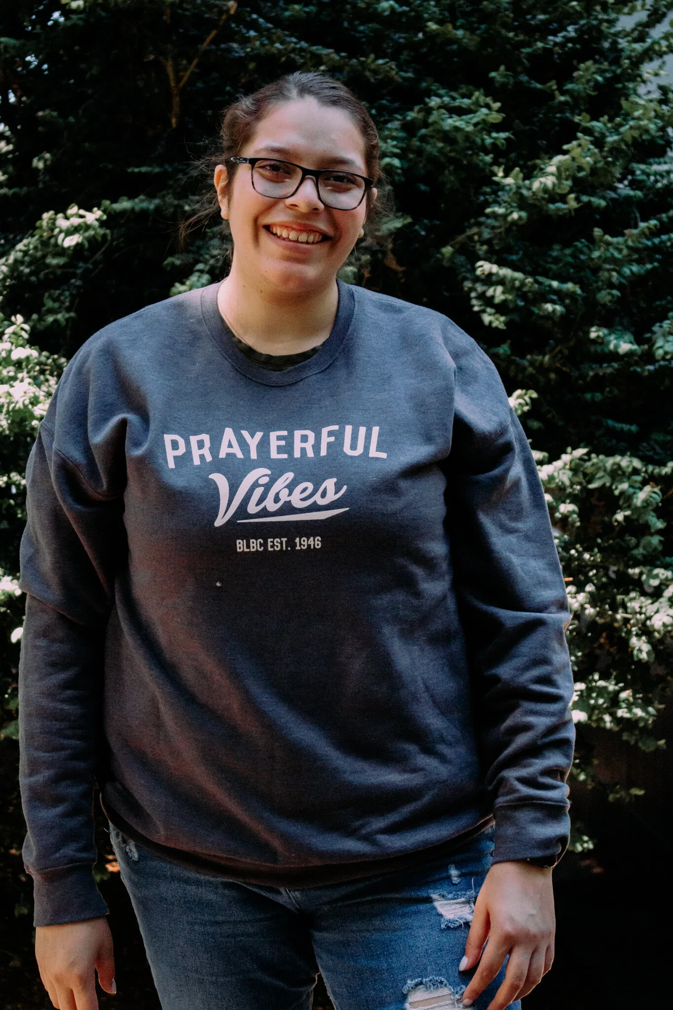 A woman with glasses and brown hair tied back, smiling outdoors in front of dark green bushes, wearing a dark gray sweatshirt with white text that says "Prayerful Vibes" and distressed jeans.