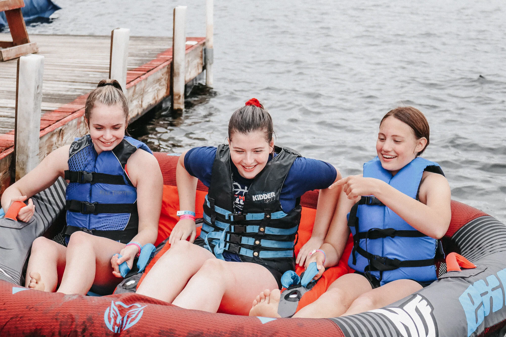Three girls wearing life jackets sitting in a boat near a dock on a body of water, smiling and having fun.