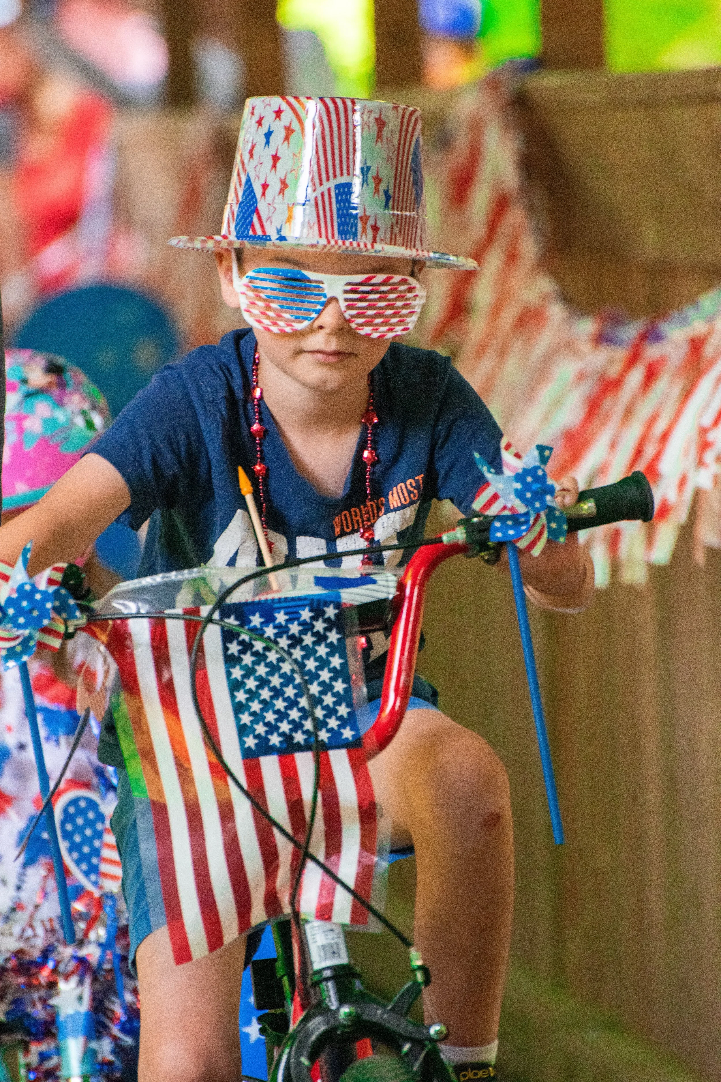 Young child wearing patriotic themed accessories, including a hat, sunglasses, and decorations on a bicycle with red, white, and blue colors and star patterns.