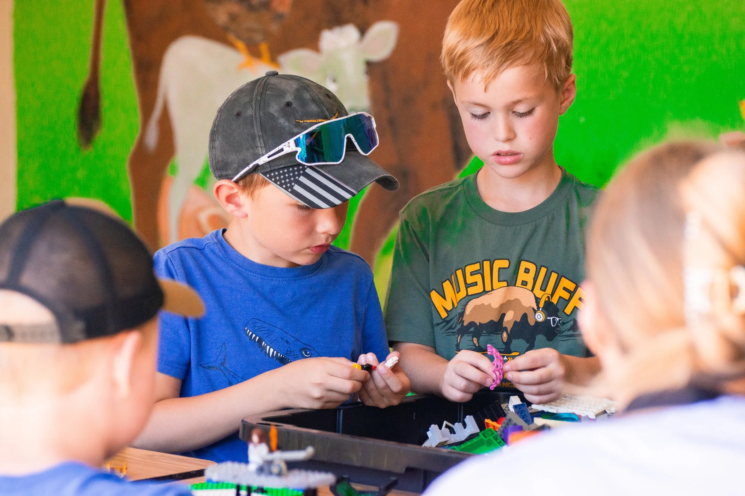 Two young boys are working with LEGO bricks at a table during a group activity. One boy is wearing a dark cap with an American flag design and blue shirt, and the other boy has red hair and a green t-shirt with a graphic print. Other children are partially visible around them, and colorful background decorations are seen behind them.