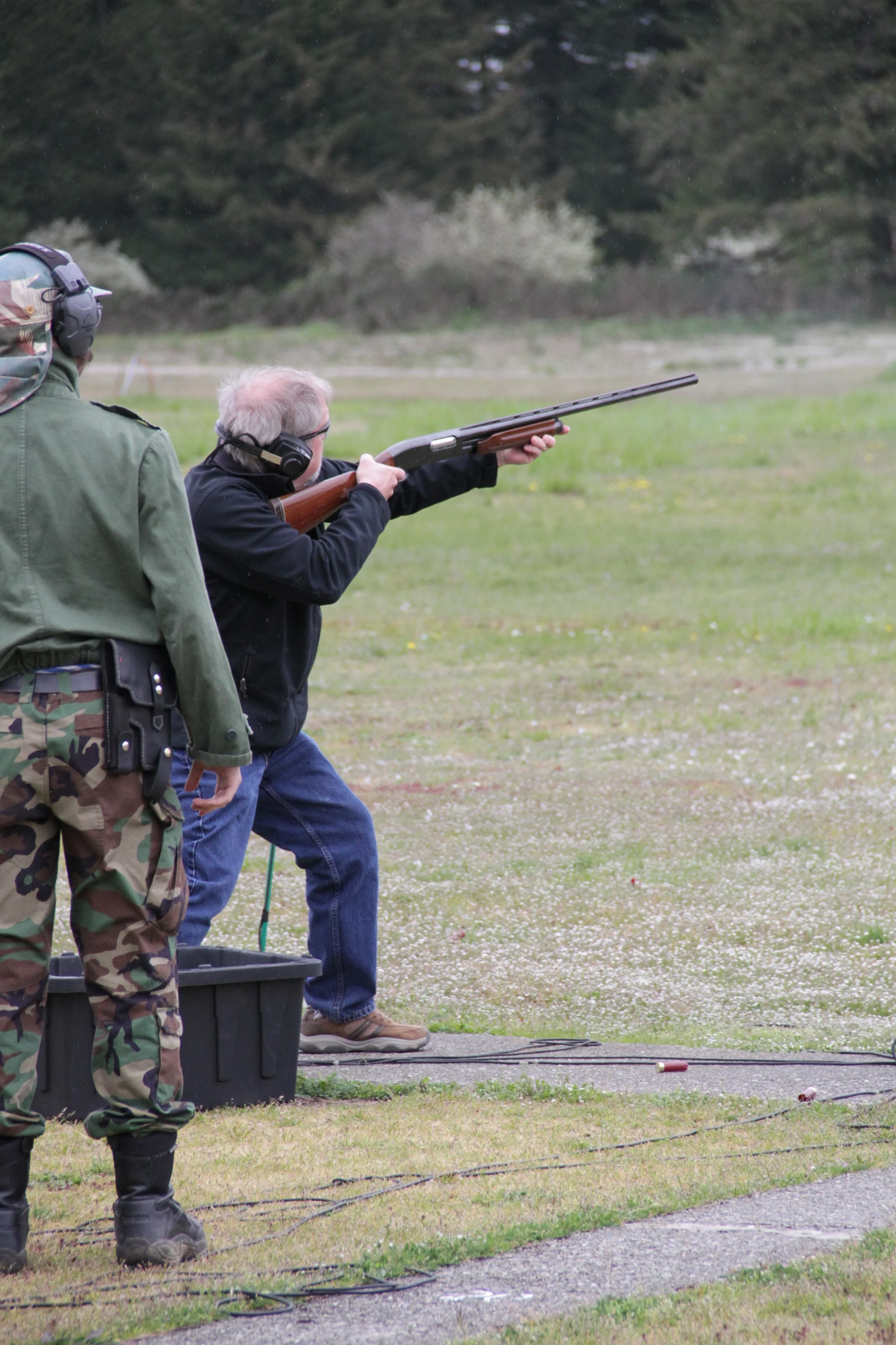 A person with gray hair and glasses is aiming and shooting a shotgun at a shooting range, with another person in camouflage standing nearby.
