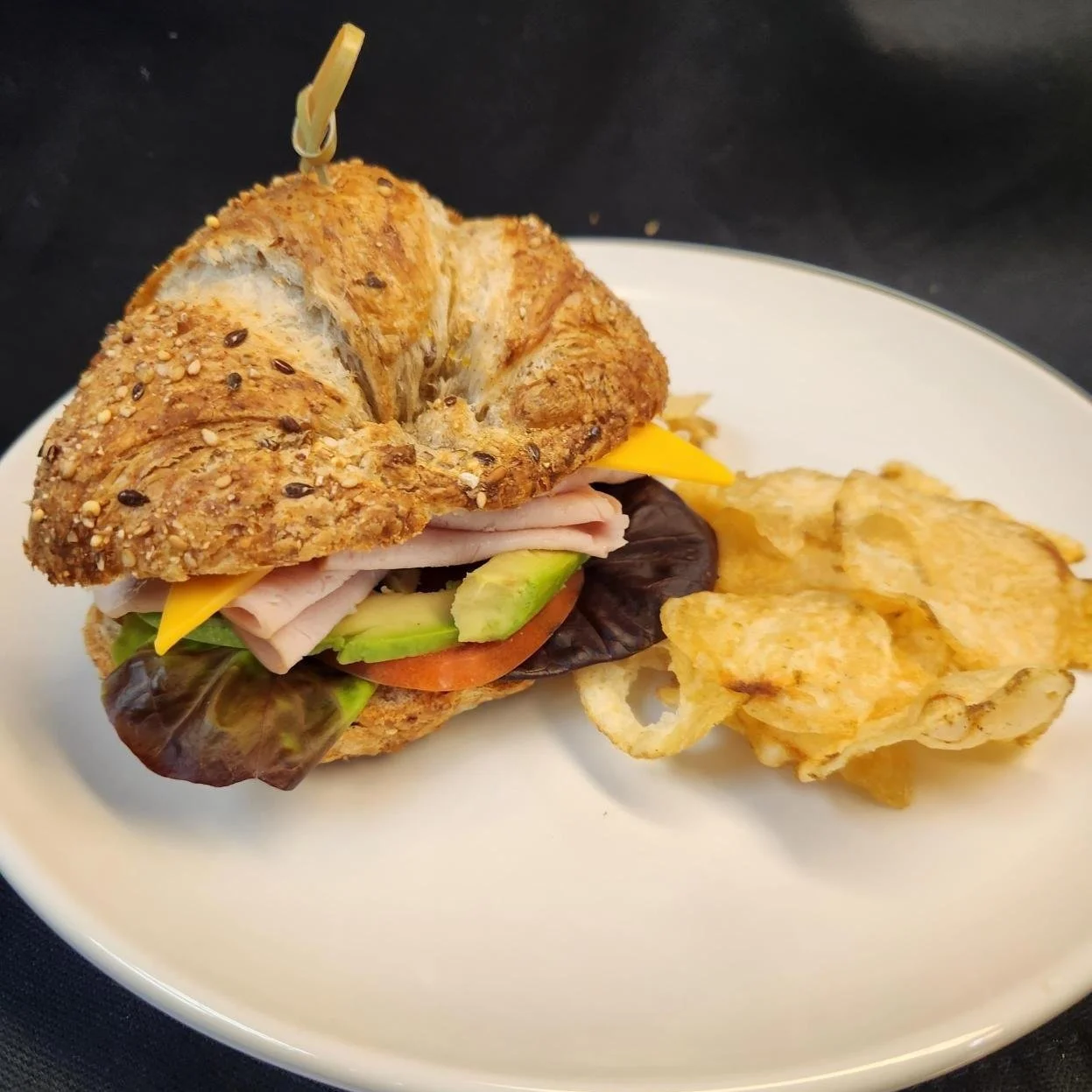 Close-up of a croissant sandwich with turkey, lettuce, tomato, cheese, and avocado, served with potato chips on a white plate.