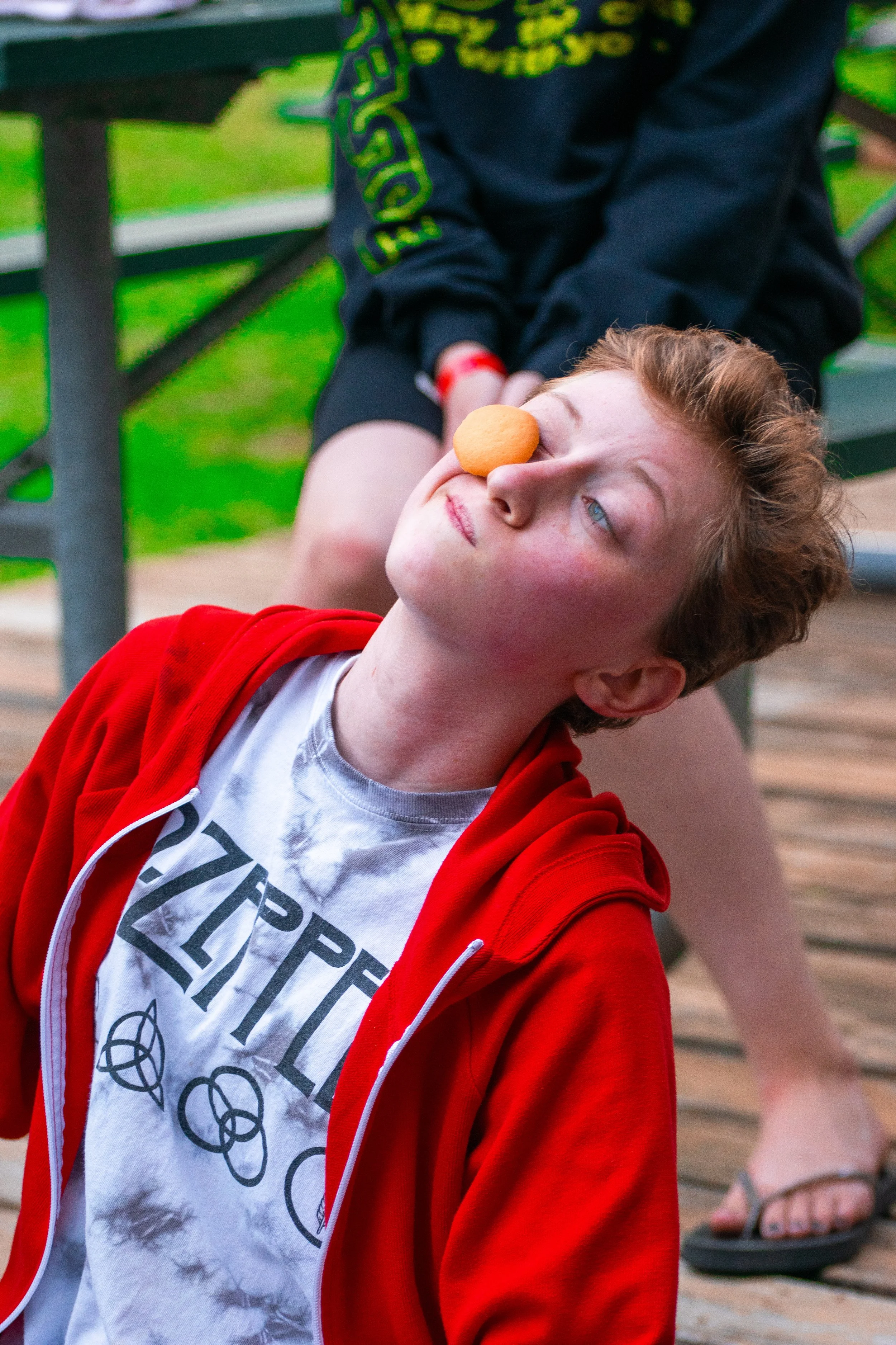 A boy with red hair and a red hoodie leaning back with a cookie on his nose in an outdoor setting with a bench and grass in the background.