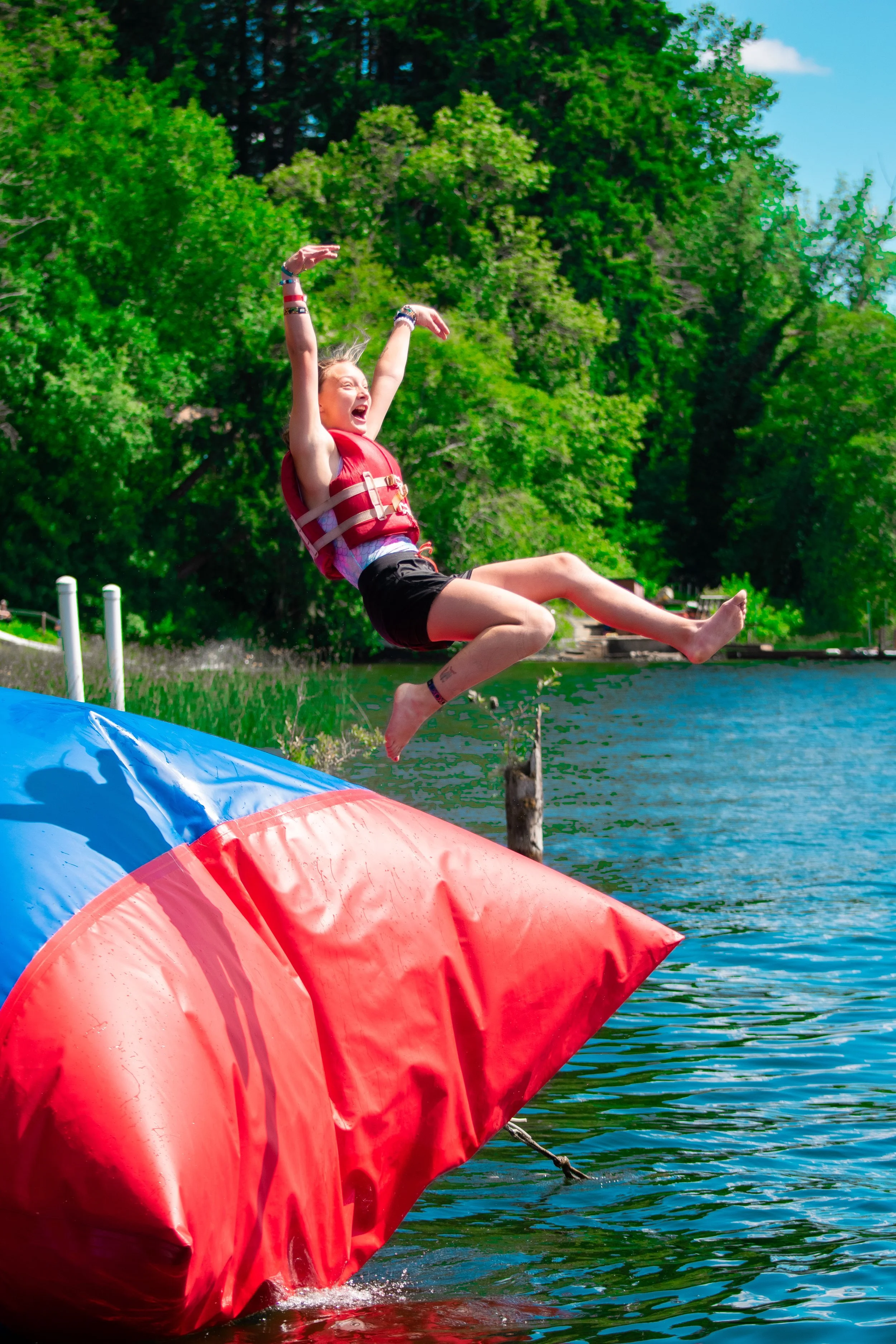 A young girl laughing and enjoying a ride on a water obstacle course with a large red inflatable part, near a lake with green trees in the background and a blue sky.