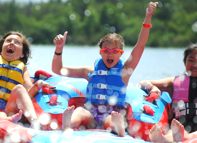 Three children on a raft in a lake wearing life jackets, smiling and celebrating.