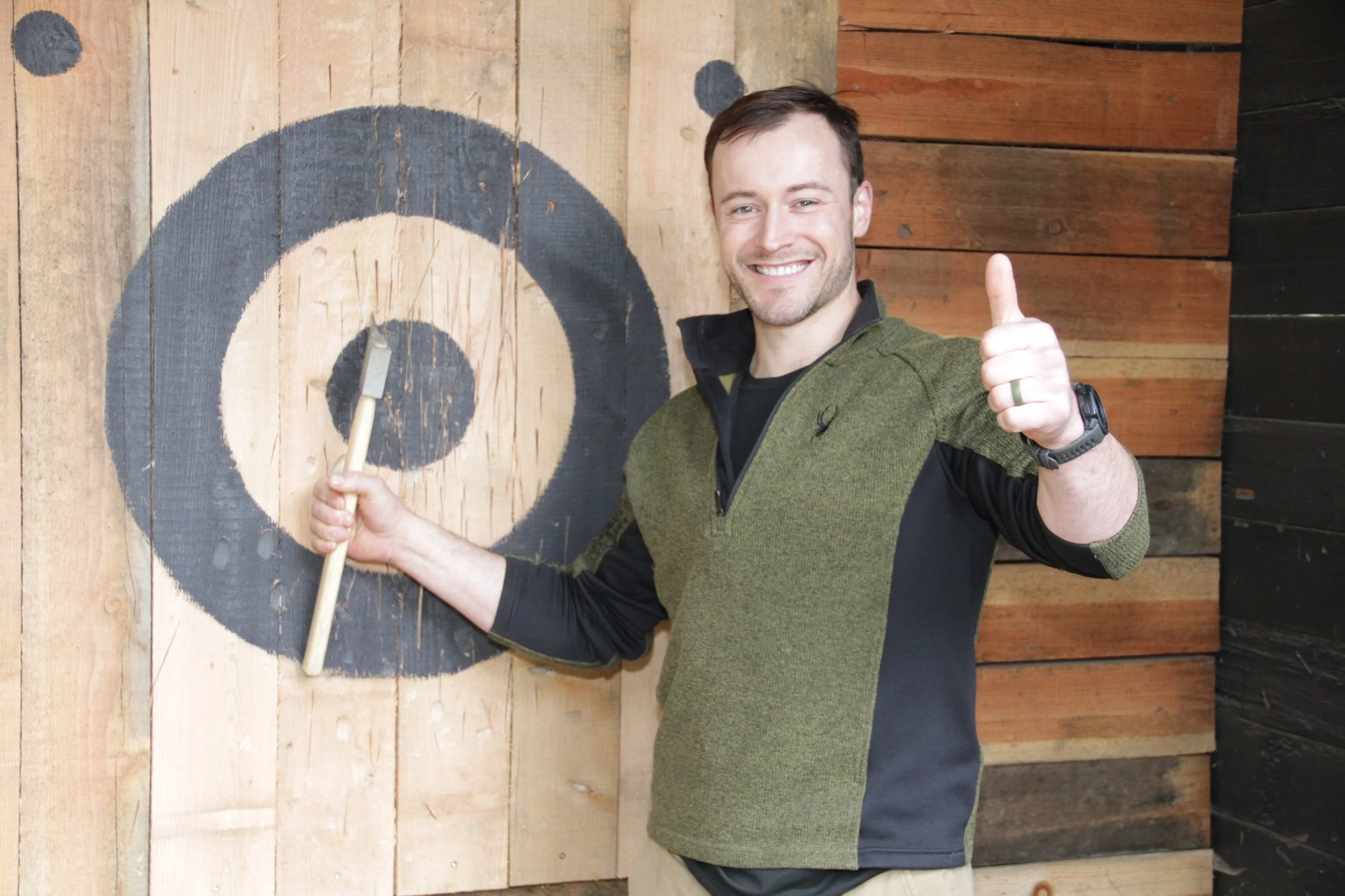 Man smiling and giving a thumbs up, standing in front of a wooden wall with a painted panda face, holding a chopstick.