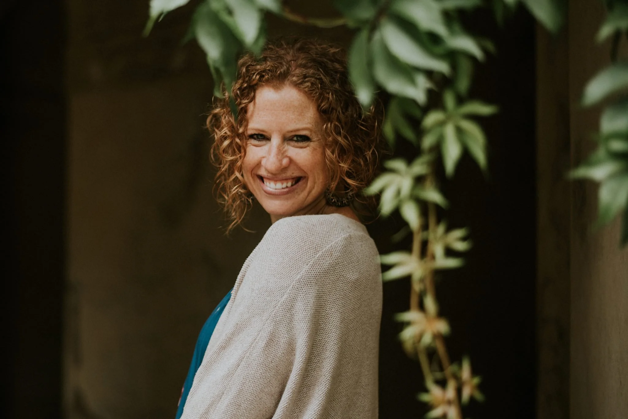 A woman with curly red hair smiling and looking over her shoulder, wearing a beige jacket and teal top, standing in front of green foliage.