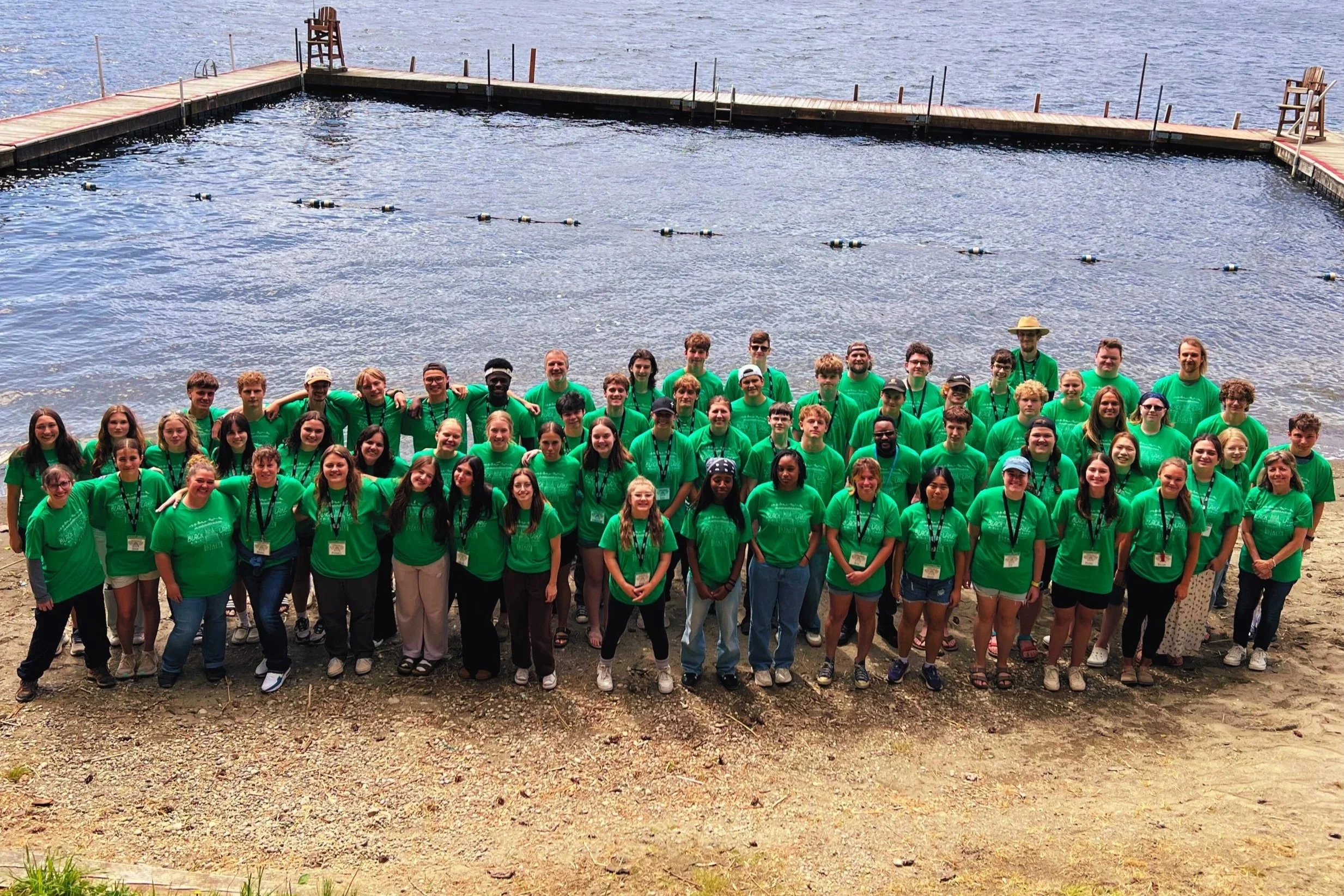 A large group of people wearing matching green T-shirts, standing outdoors near a lake or river, with a wooden dock and chairs in the background.