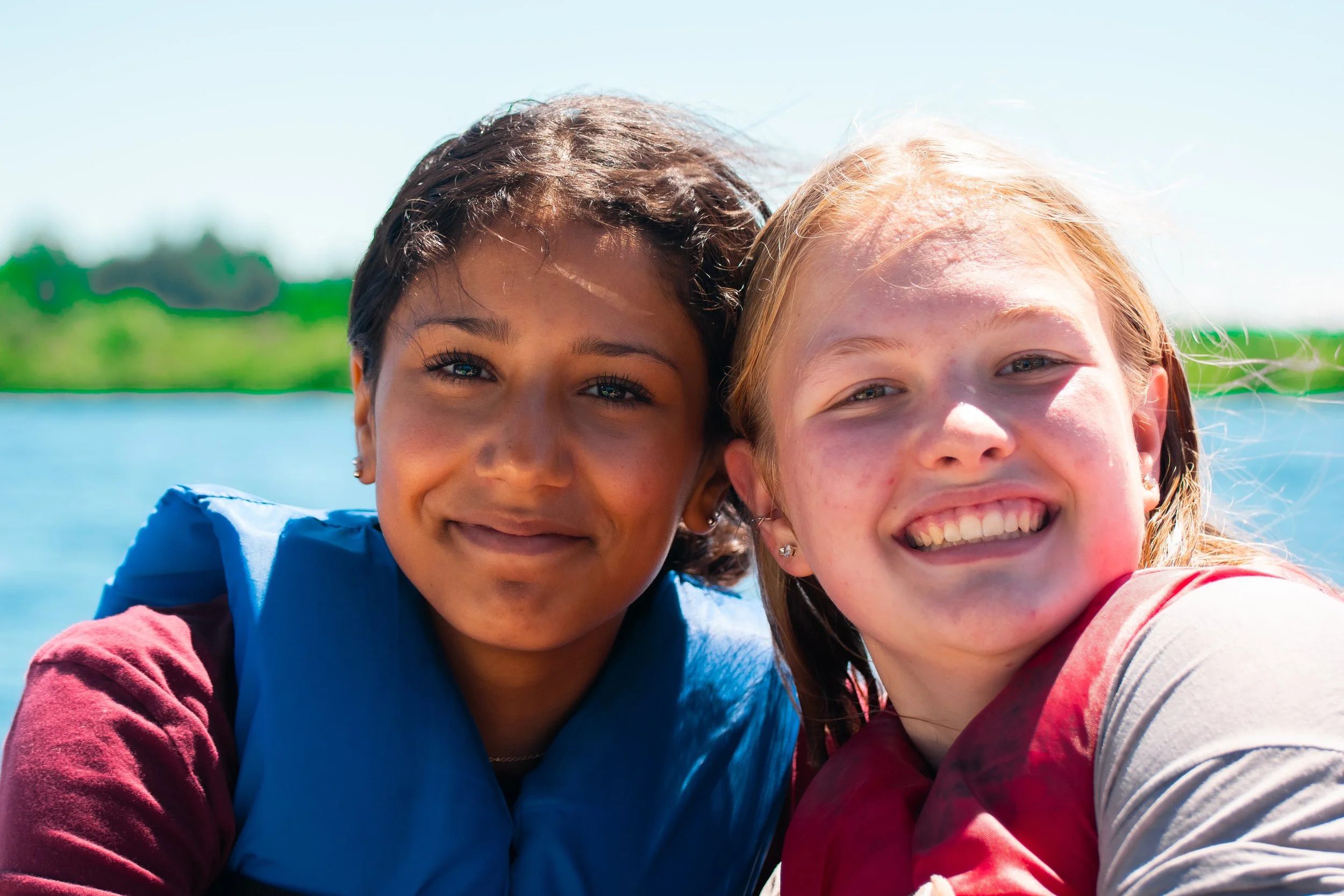 Two smiling girls with arms around each other outdoors on a sunny day, near water with green trees in the background.