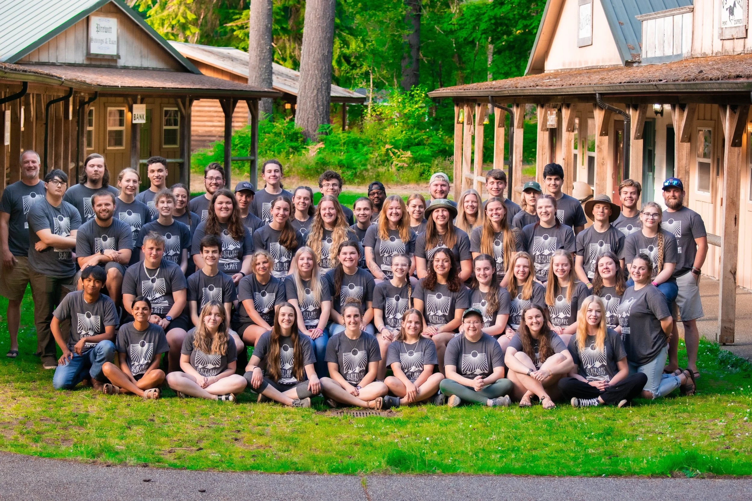 A large group of people, mostly young adults, posing outdoors in front of wooden cabins in a wooded area with tall trees and green foliage. Many are wearing matching gray T-shirts with a design on the front.
