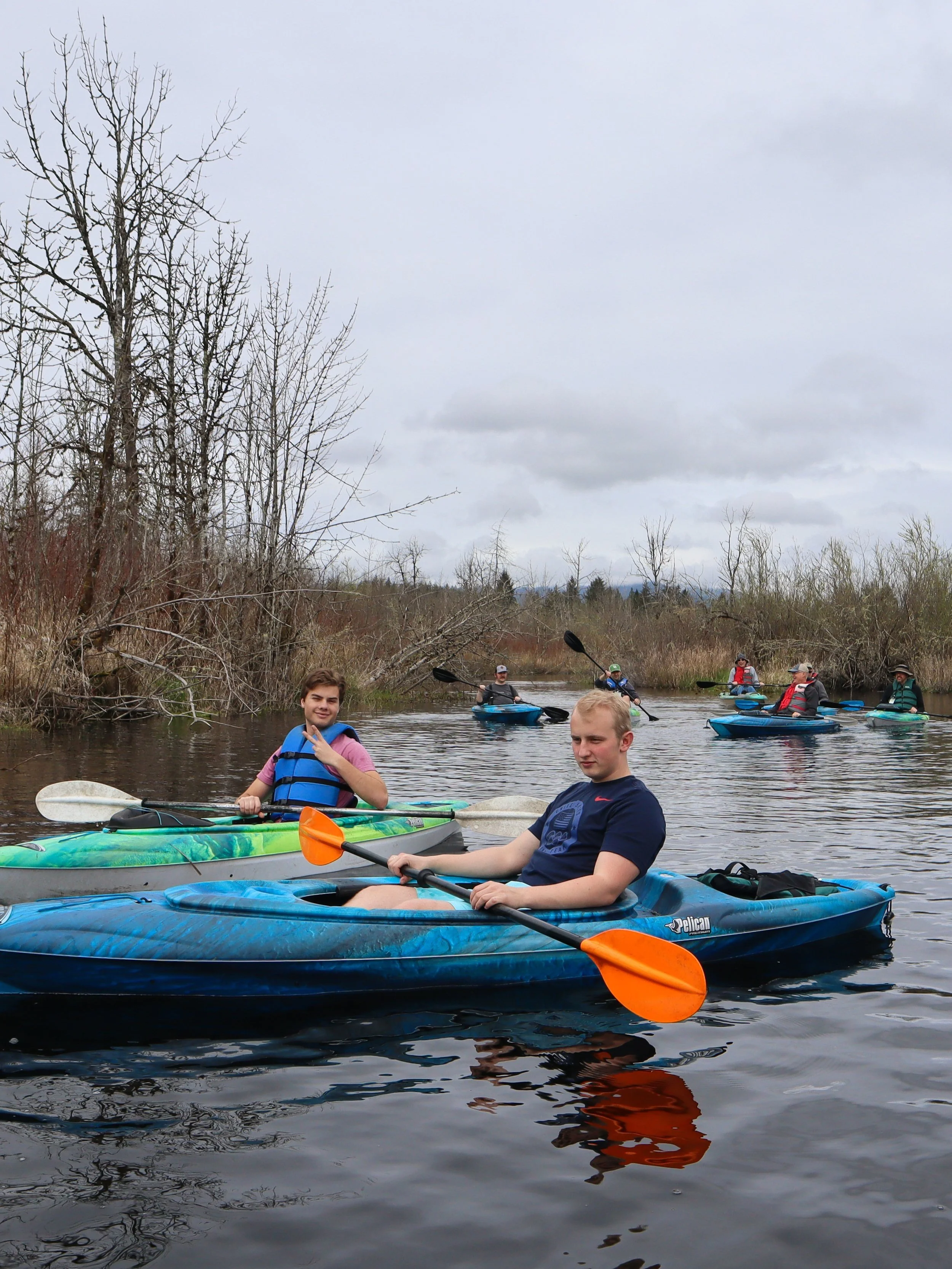 A group of people kayaking on a calm river. The sky is overcast with gray clouds, and there are leafless trees along the riverbanks. Two young men are in the foreground, one smiling and wearing a blue life jacket, and the other looking down, both in kayaks. Several others are paddling in the background.