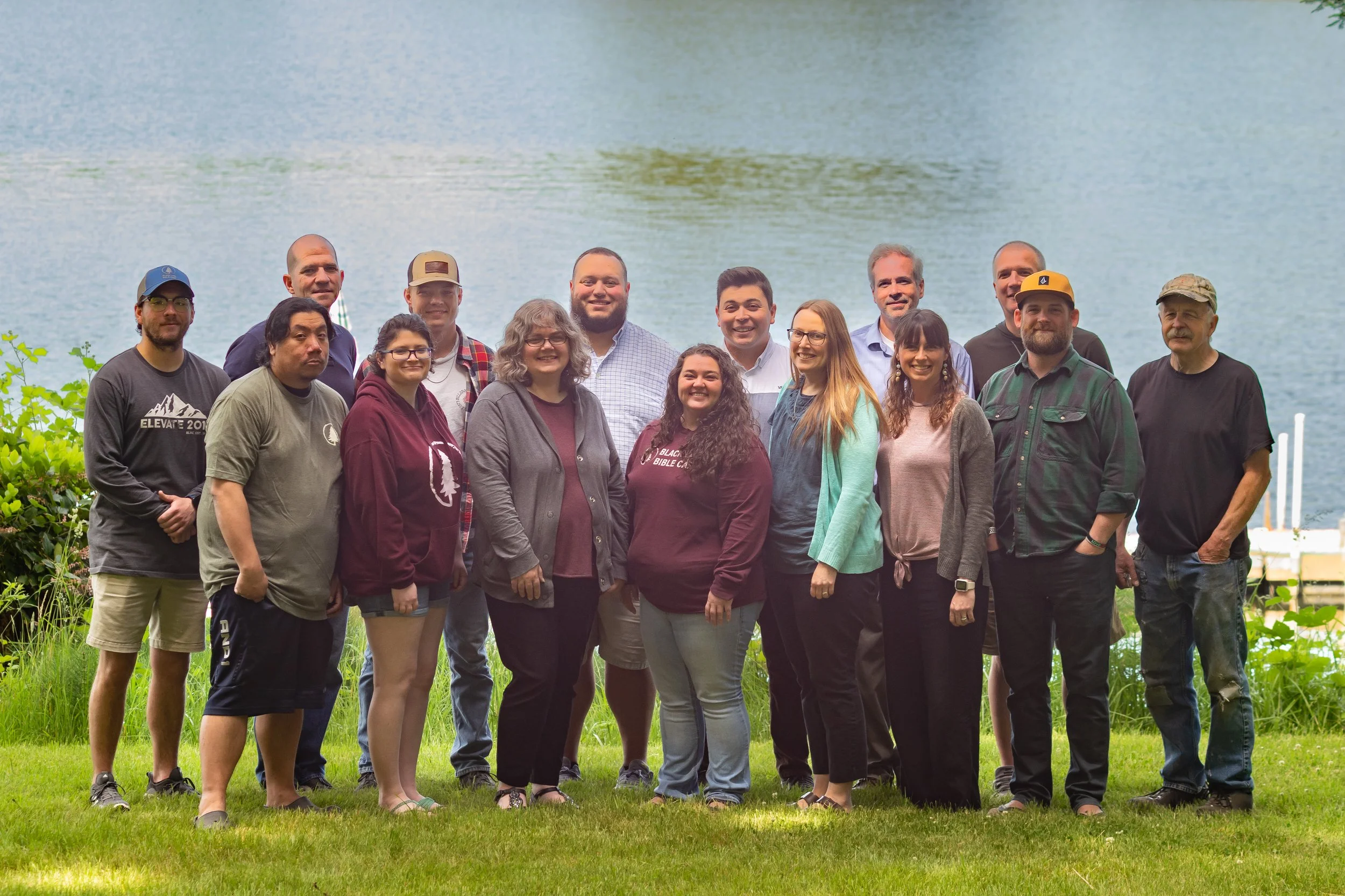 Group of fifteen people standing outdoors on grass with a lake in the background, smiling at the camera.