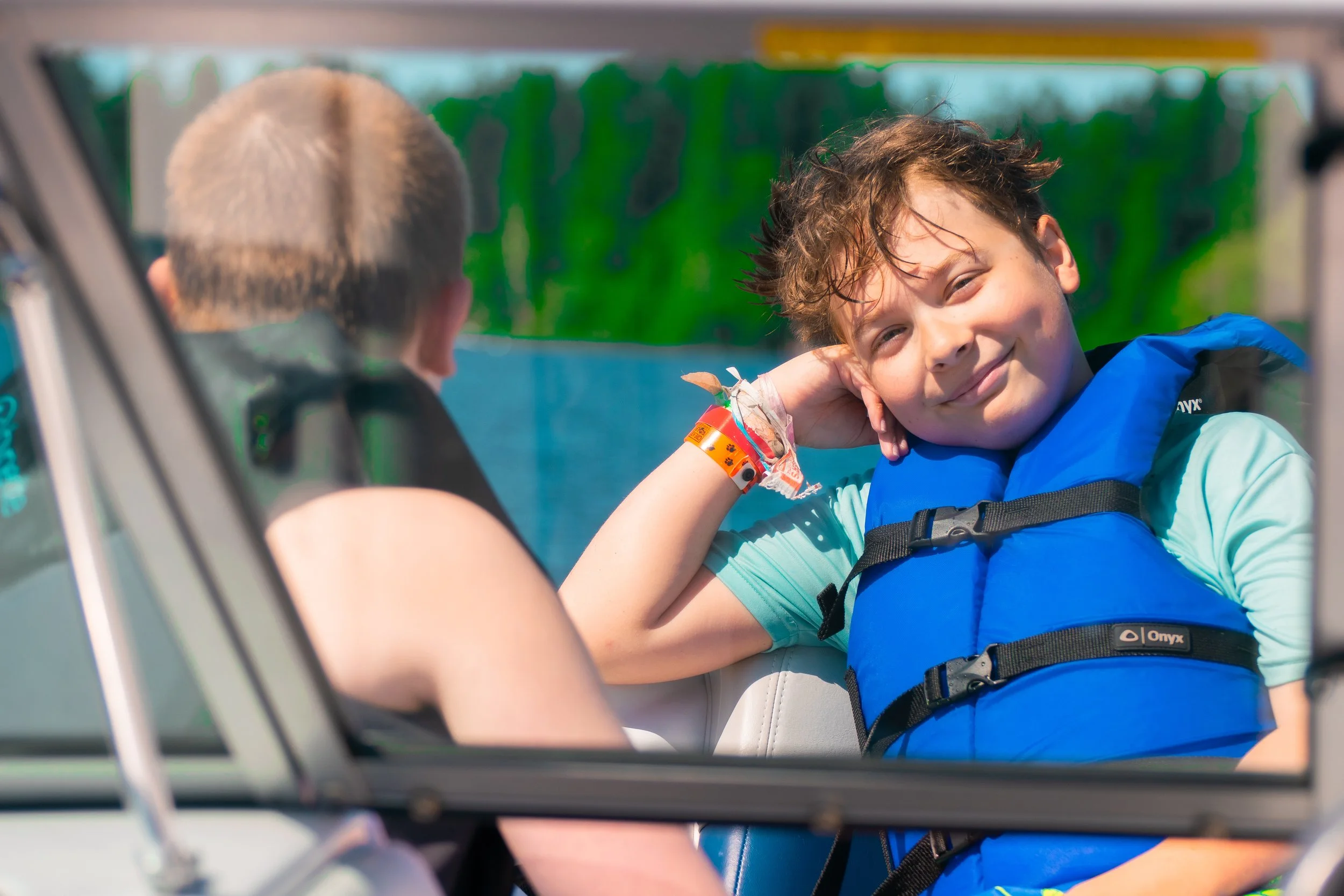 Young boy with messy hair smiling and leaning his head on his hand, sitting in a boat with a life jacket, facing an adult with short blonde hair inside a boat, water and greenery in the background.
