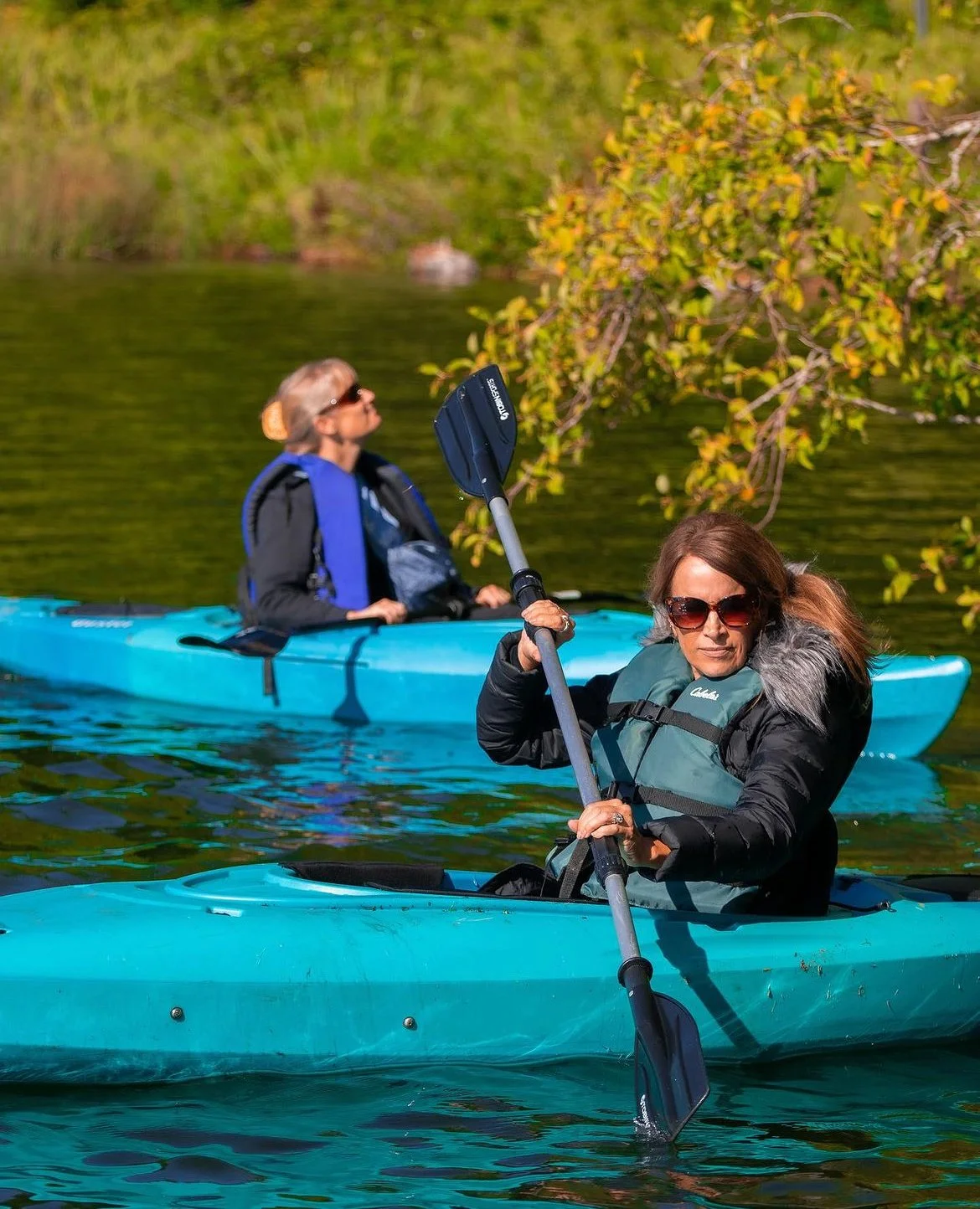 Two women kayaking on a calm river, surrounded by green trees and foliage, each wearing life jackets and sunglasses, with one woman in the foreground holding a paddle.