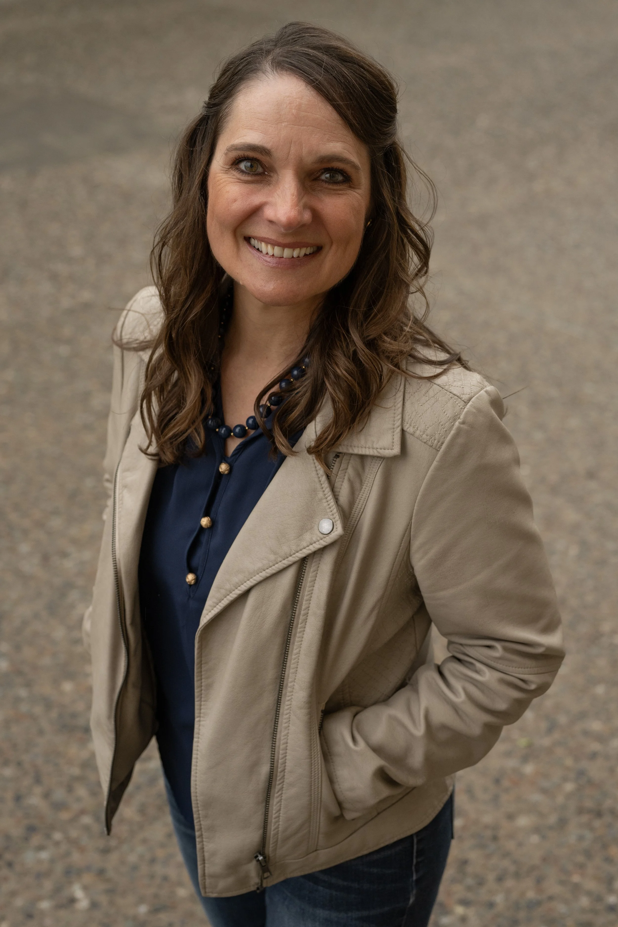 A woman with brown hair and green eyes smiling, wearing a beige jacket, navy shirt, and a blue beaded necklace, standing outdoors on a gravel path.