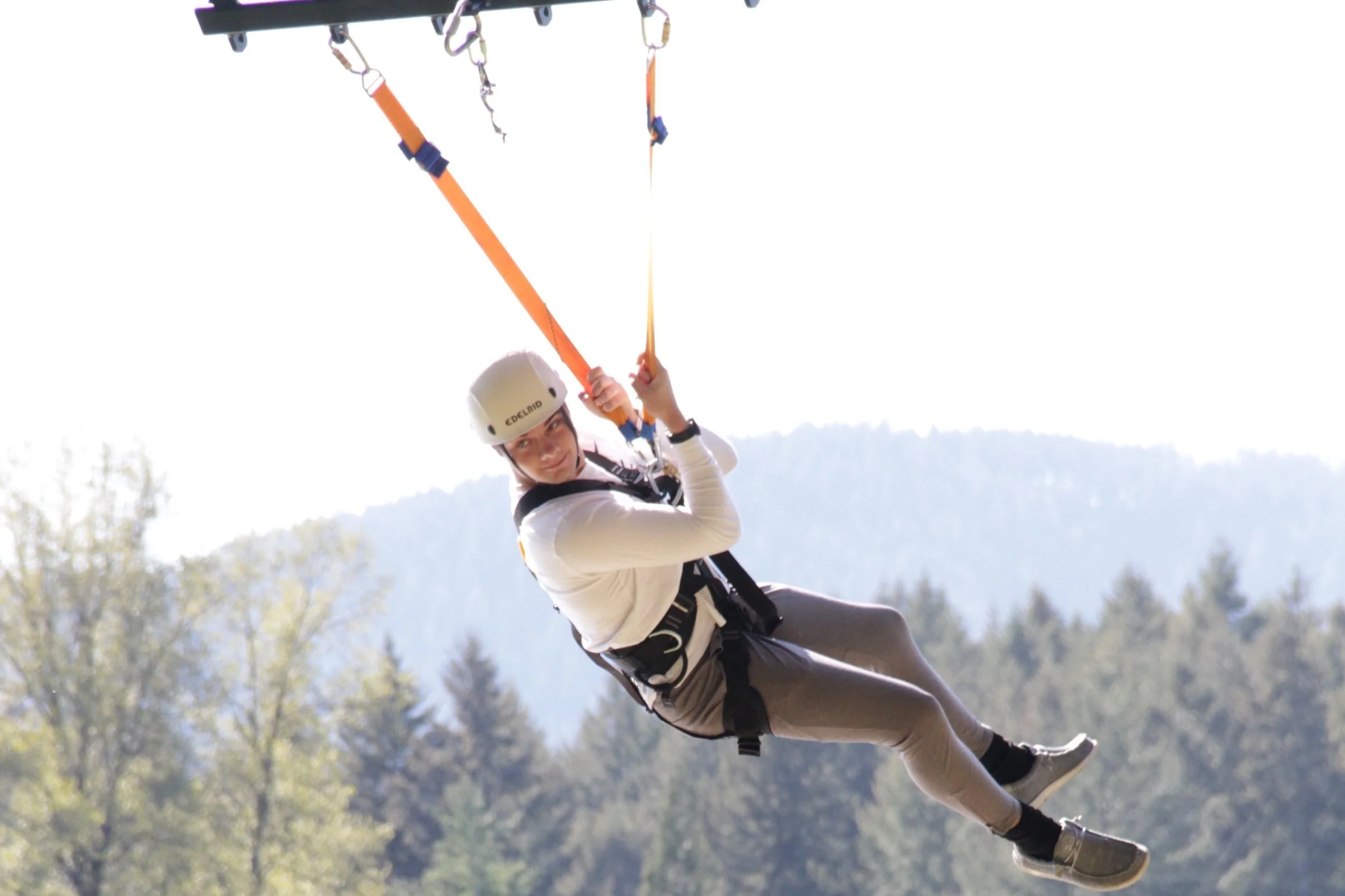 A person, wearing a helmet and harness, is ziplining through a forested area with trees and mountains in the background.