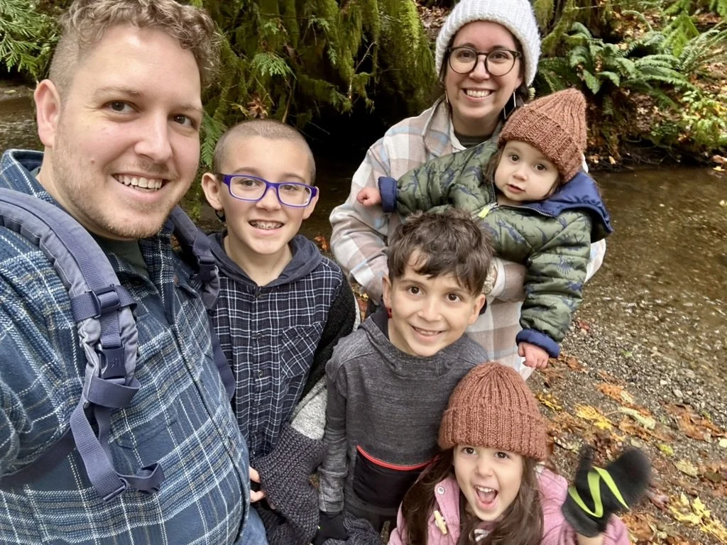 A family of six outdoors in a forest near a creek, taking a selfie. The group includes two adults and four children, all dressed in casual outdoor clothing and warm hats. Leafy trees and moss are visible in the background.
