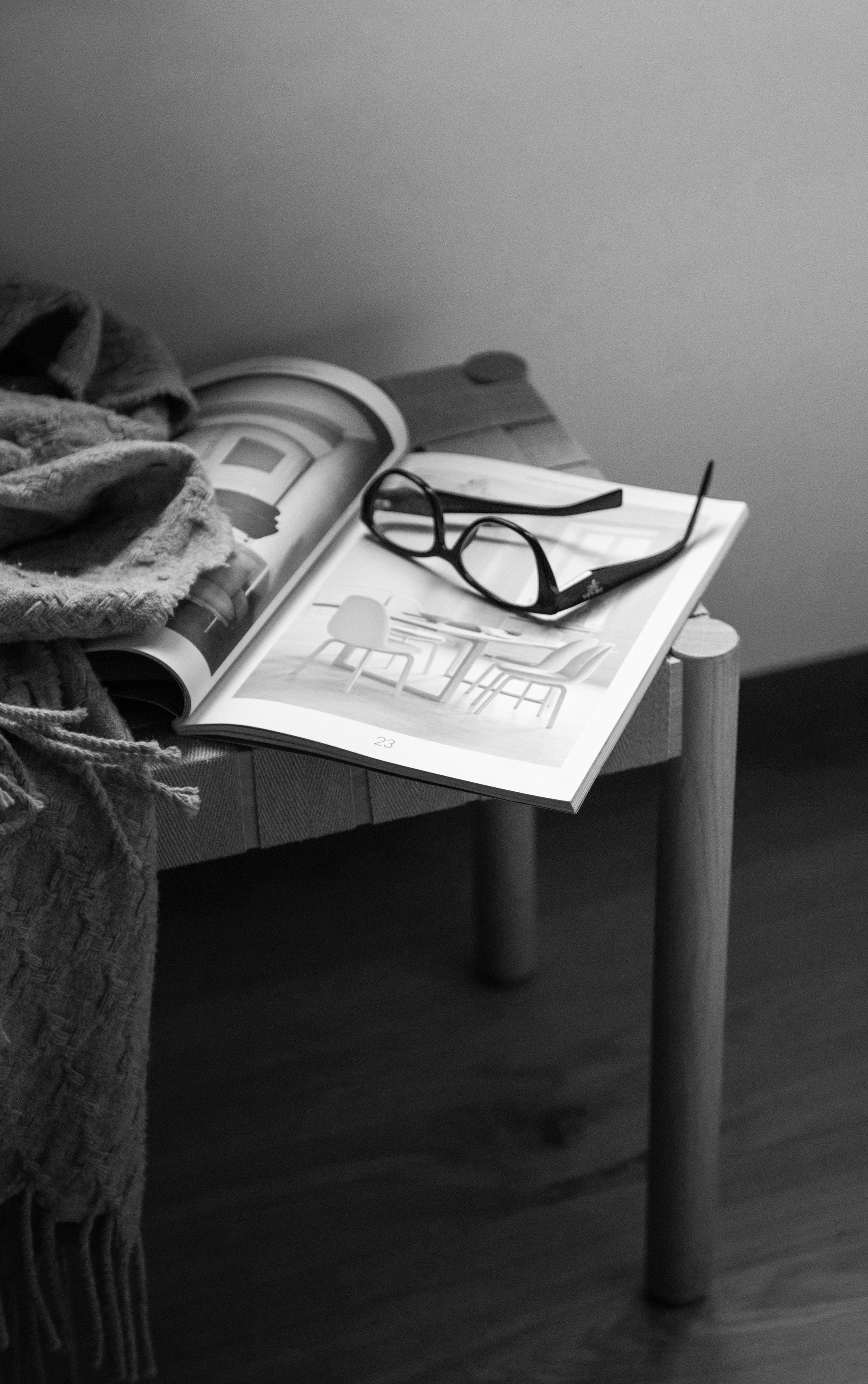 A black and white photo of a small table with a pair of glasses, an open magazine, and some clothing or fabric items on the edge of the table. Katie McFarlan Dakota Design Company best templates for interior designers