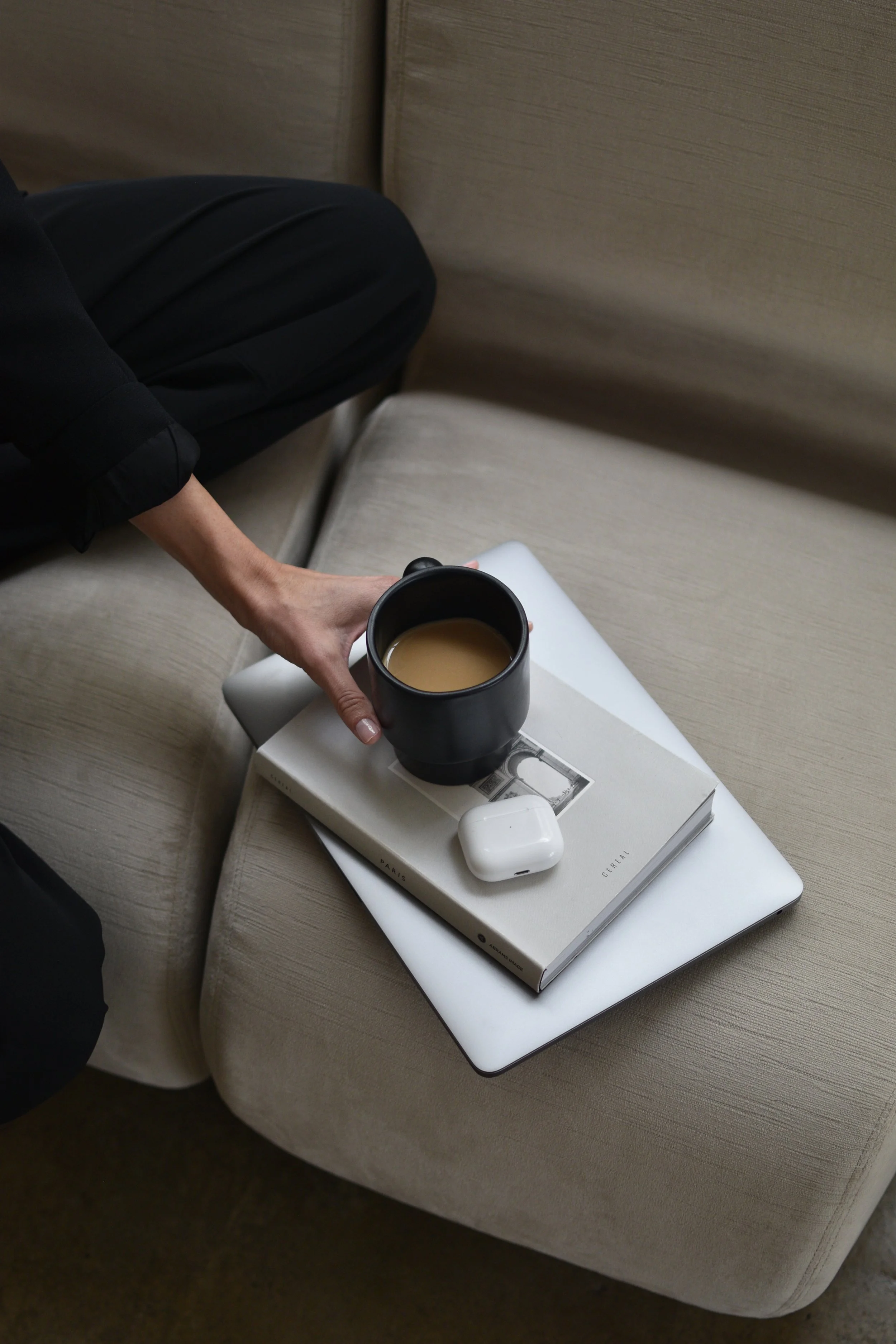 A person's hand holding a black mug of coffee or tea on a beige sofa, next to a closed white laptop, a hardcover book, and white wireless earbuds.Katie McFarlan Dakota Design Company Best Client Process Templates for Interior Designers