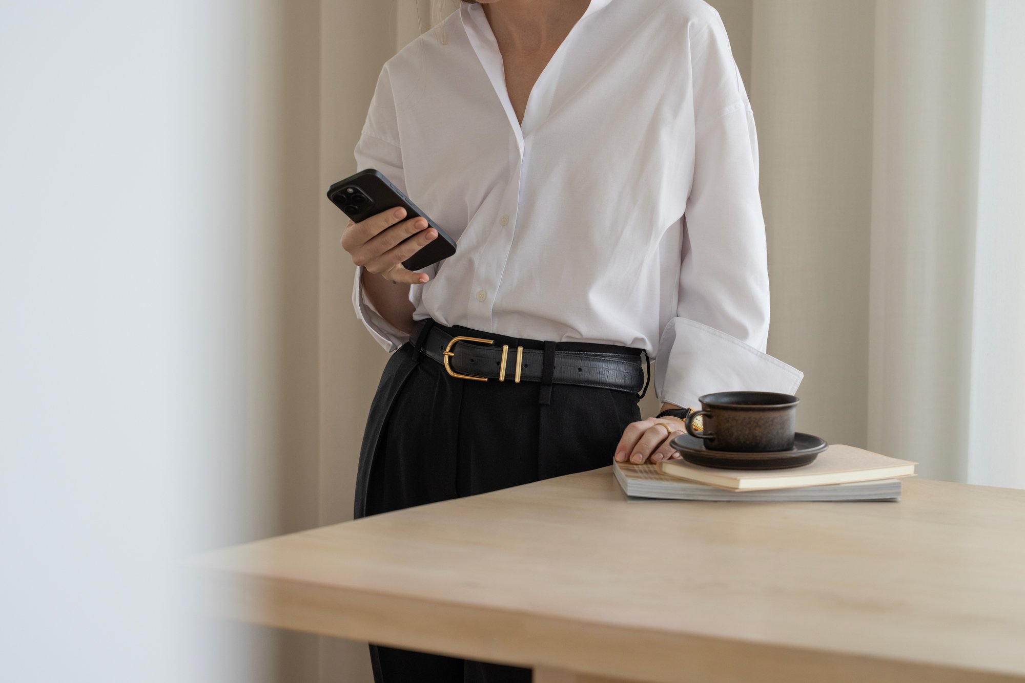 A woman wearing a white shirt and black pants with a black belt, holding a smartphone.Katie McFarlan Dakota Design Company Best Client Process Templates for Interior Designers