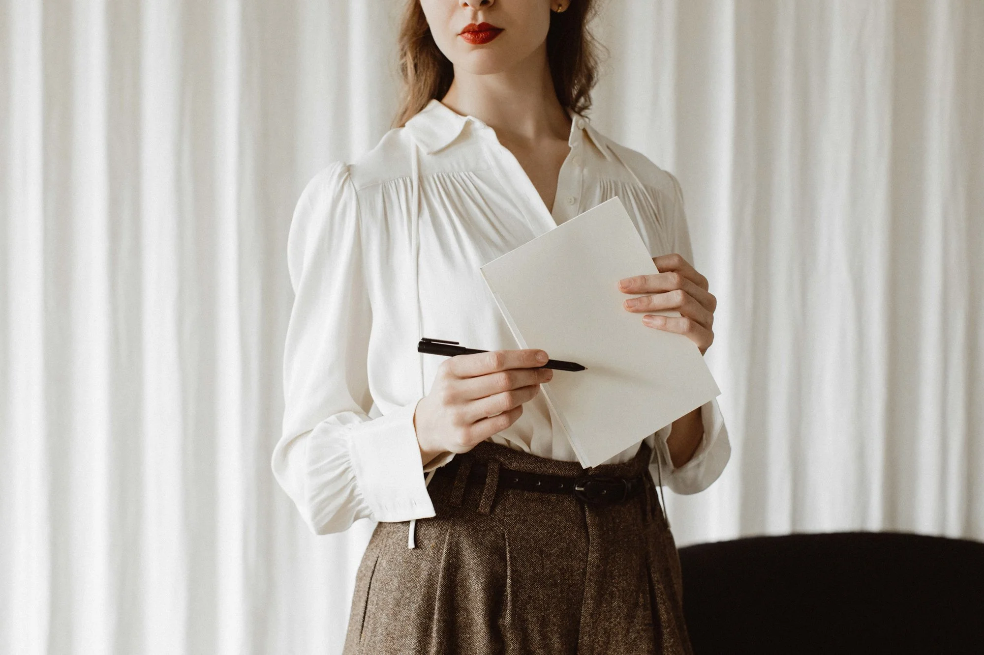 A woman with red lipstick wearing a white blouse and brown high-waisted trousers holding a blank notebook and pen.Katie McFarlan Dakota Design Company Best Client Process Templates for Interior Designers