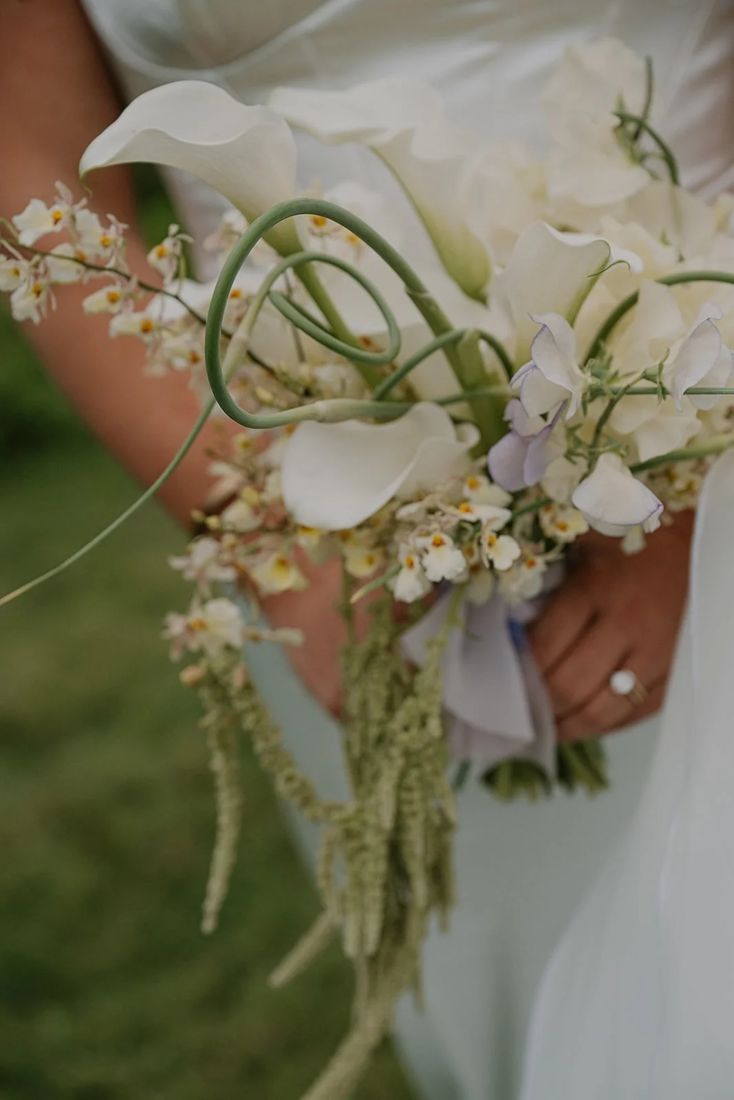 A close up of a modern white bridal bouquet with calla lilies, garlic scapes, sweet pea, amaranth and orchids