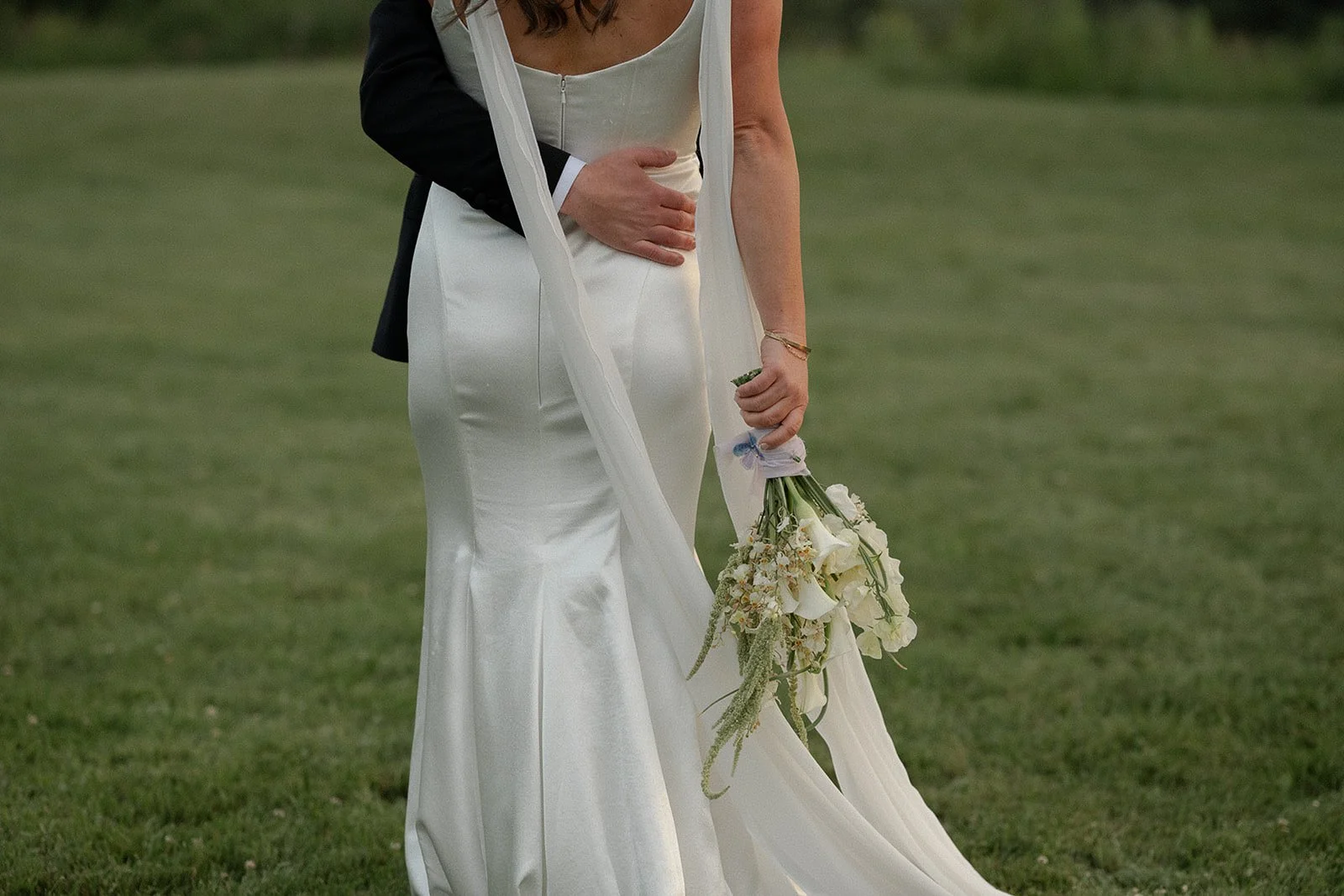 A bride holding a bridal bouquet of white calla lilies, garlic scapes, oncidium orchids and green amaranthus embraces her groom at Mt. Hood Organic Farms.