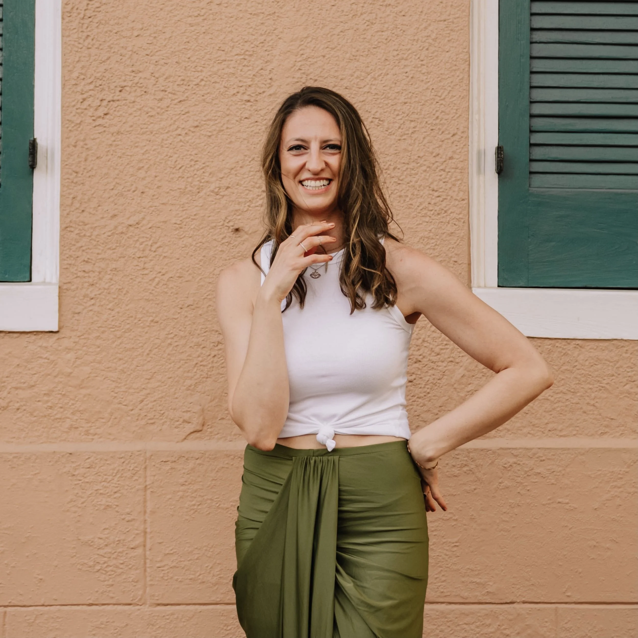 Woman smiling in white top and green skirt against a peach textured wall with green shutters.