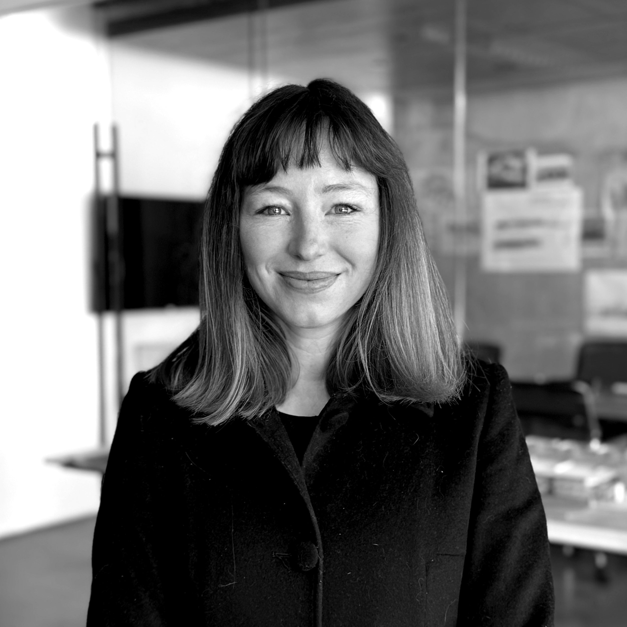 A woman with shoulder-length hair and bangs smiling at the camera in an indoor setting with blurred background of office furniture.