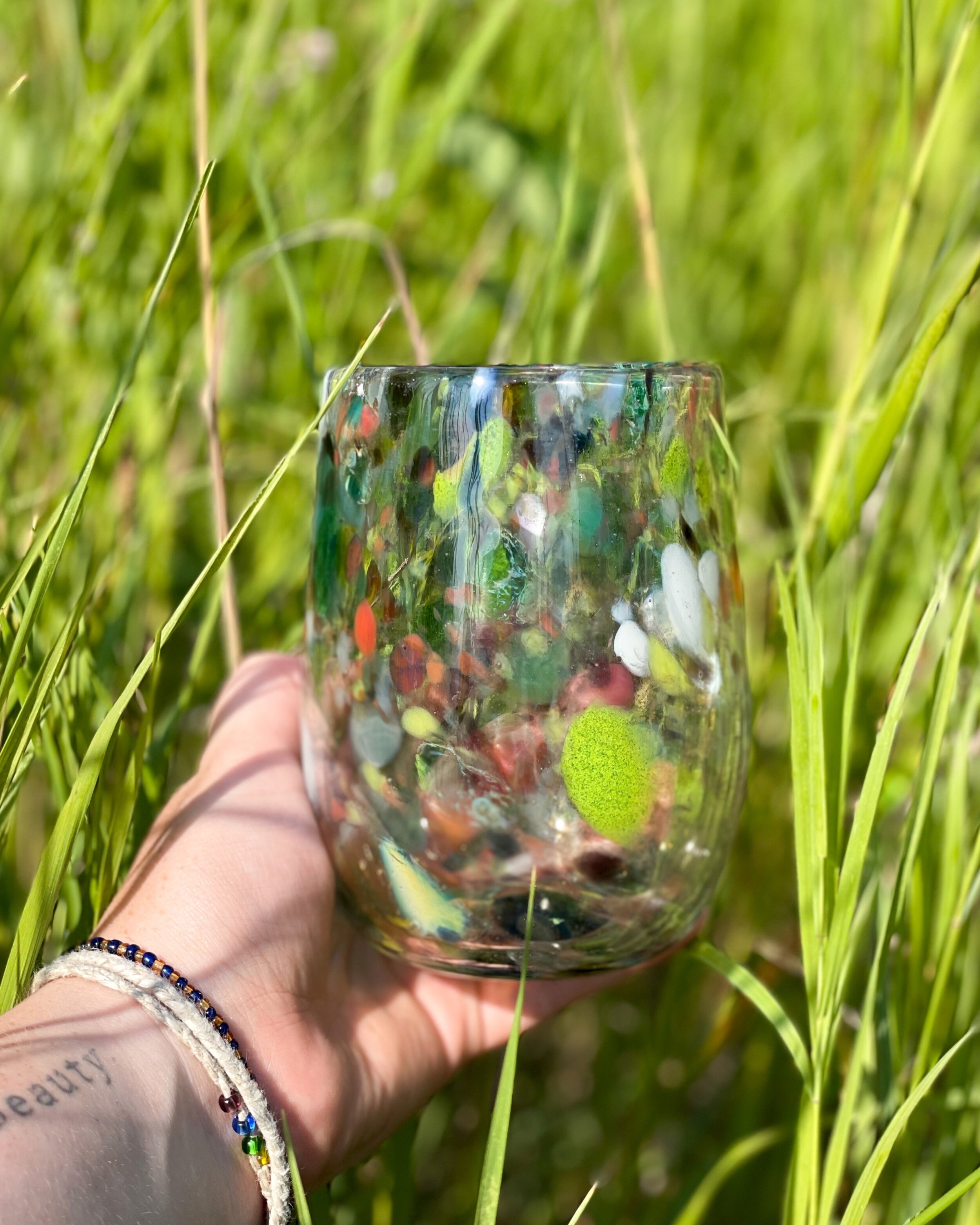 Blown glass stemless wine glass - speckled with many colours - held by a hand in tall grass