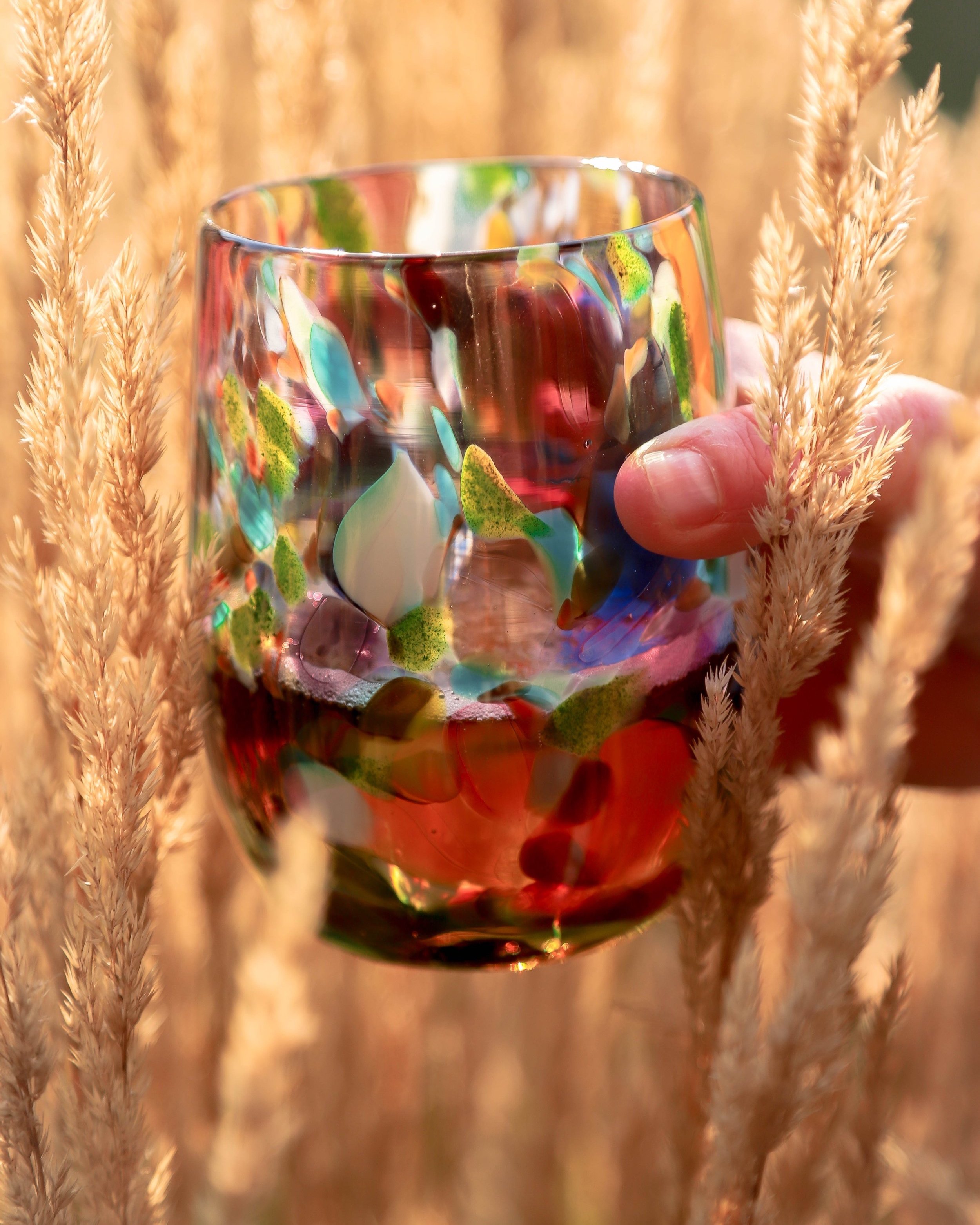 Blown glass stemless wine glass - speckled with many colours - held by a hand in tall wheat type grass
