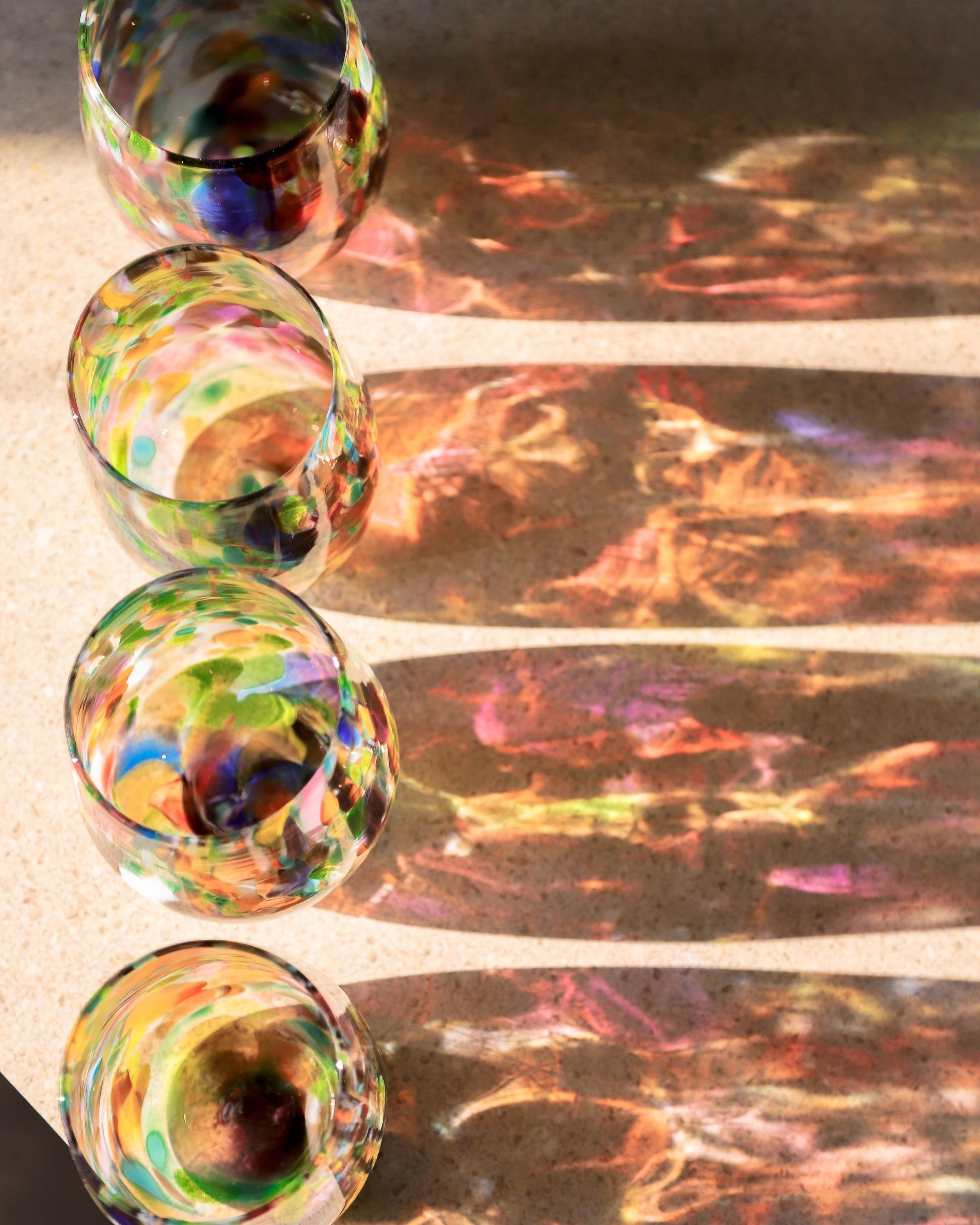 4 bllown glass stemless wine glasses - speckled with many colours - with sun shining through the cups with a reflection on the counter