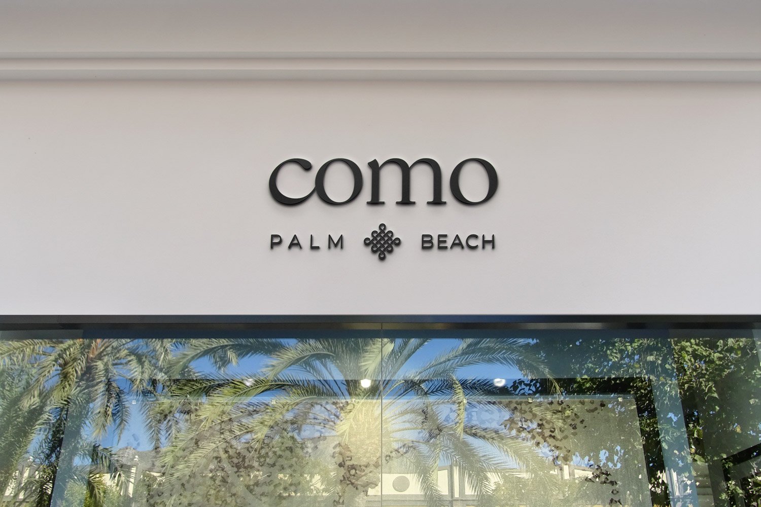 Shop front with the sign 'como Palm Beach' above a large glass window reflecting palm trees and blue sky.