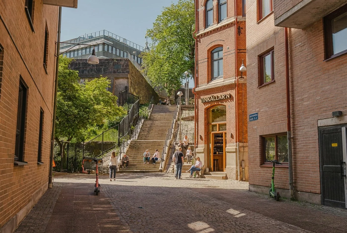 Gata med trappor som leder upp till en park, omgiven av bygghus i historisk stil, med människor som sitter och går. Grön träd, tydligt blå himmel, i Stockholm, Sverige.