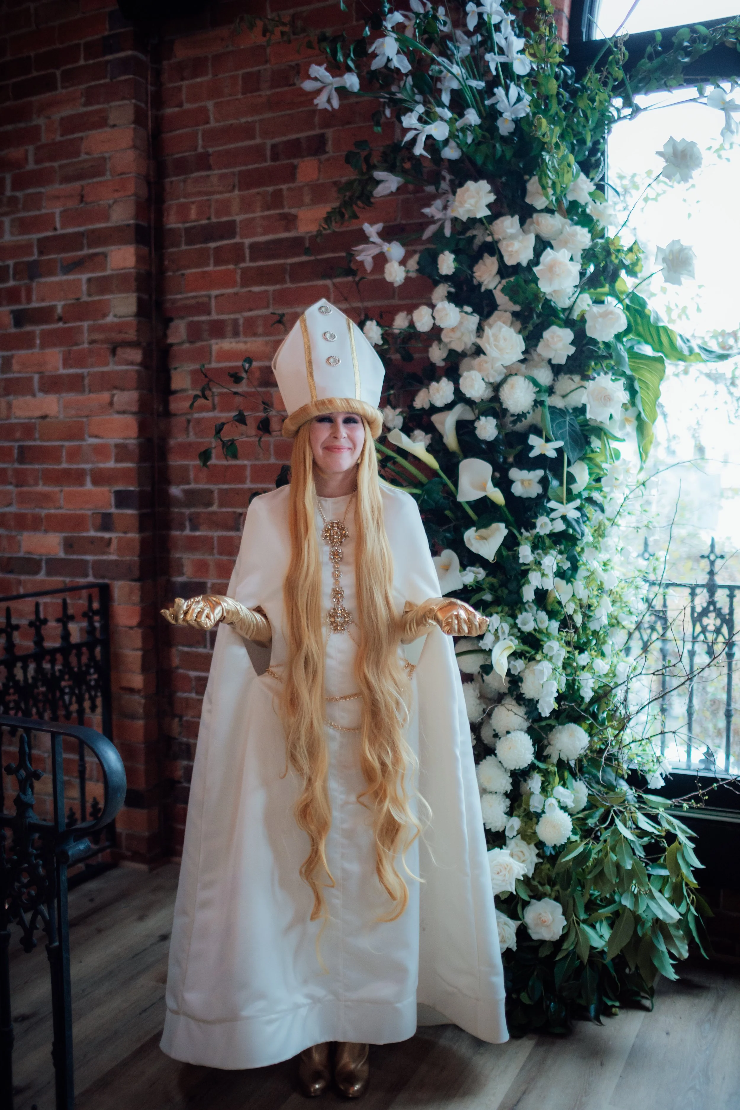 A person dressed as a royal figure or king, wearing a white gown and gold gloves, standing in front of a brick wall and white floral decorations, with a large ornate hat on their head.