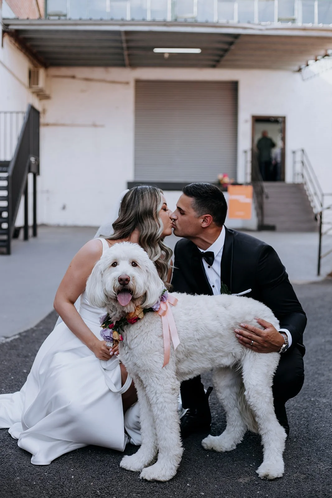 A bride and groom, dressed in wedding attire, kiss while kneeling outside a building with a large dog in front of them. The dog has white, curly fur, is wearing a floral collar with ribbons, and looks happy with its tongue out.