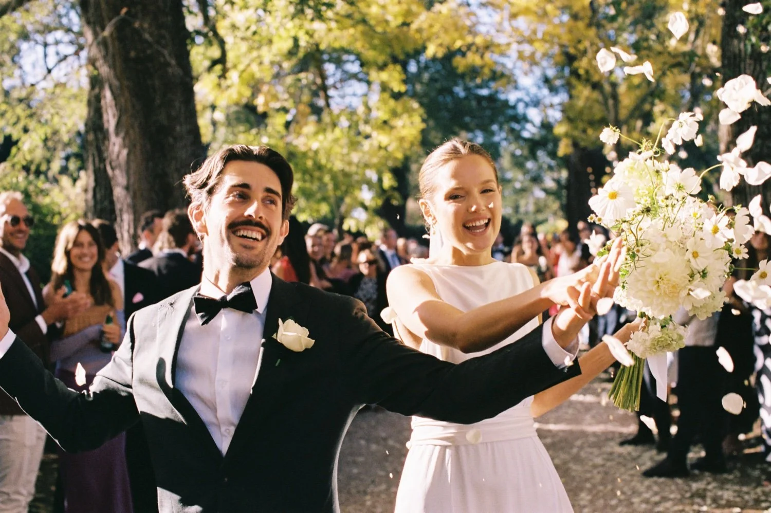 A joyful couple at their outdoor wedding reception, surrounded by guests and trees, throwing flower petals.