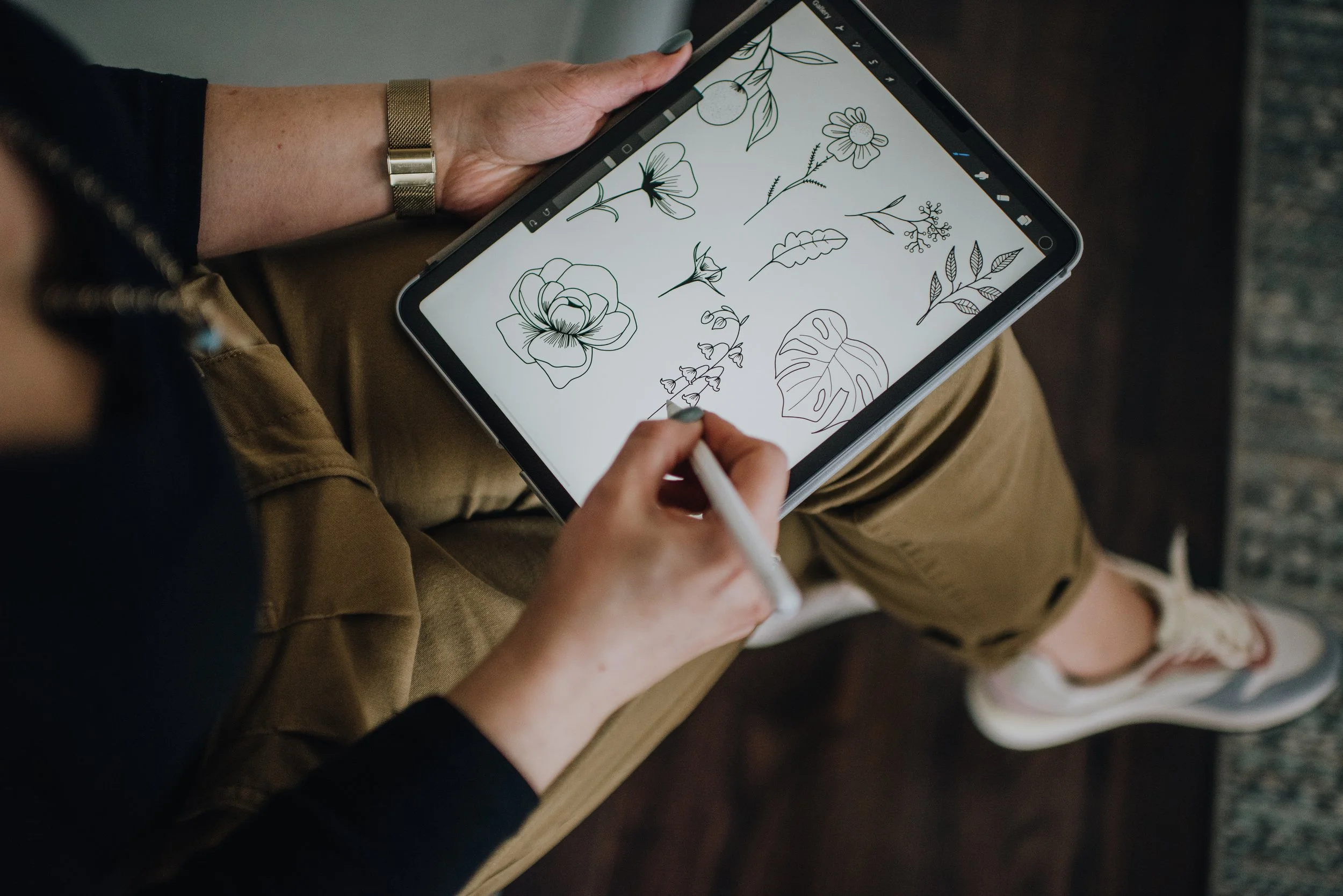 Mari McCallion drawing floral illustrations on a digital tablet with a stylus, sitting on a wooden floor.