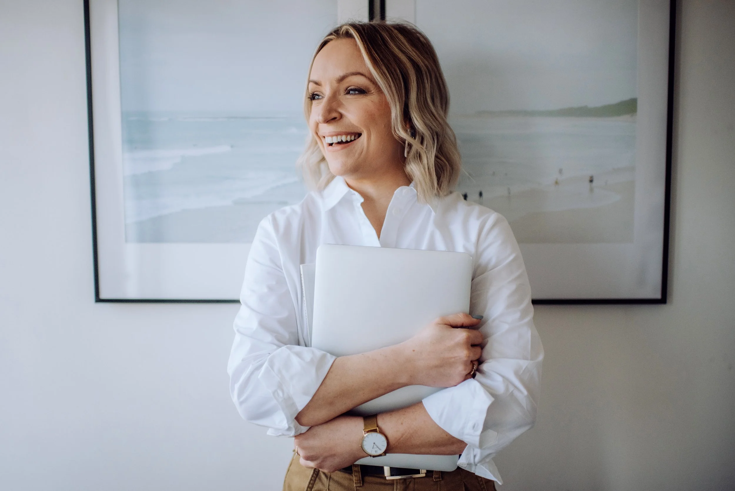 Mari McCallion smiling and holding a closed laptop, standing in front of a framed beach photograph on the wall.