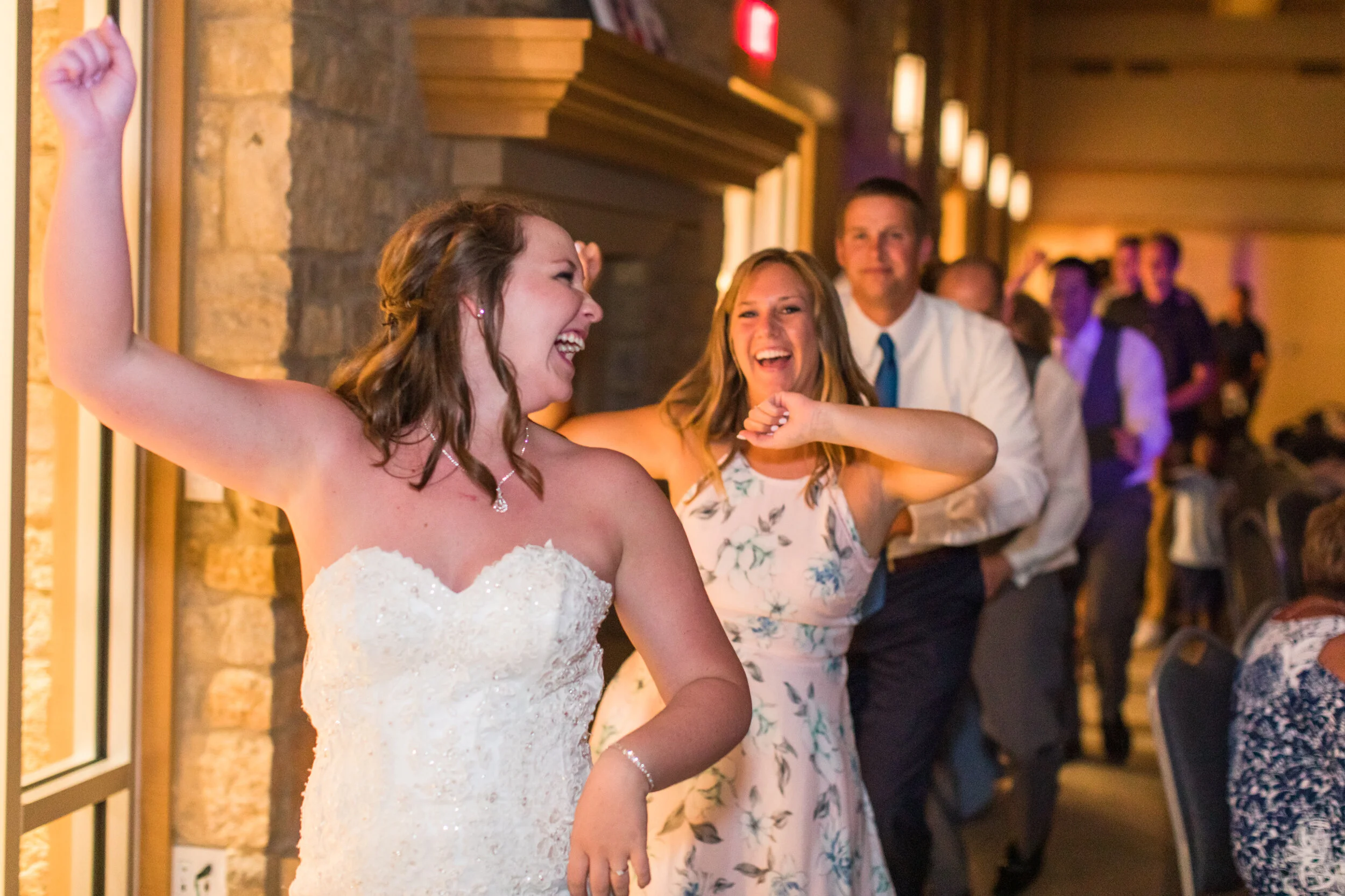 A woman in a white wedding dress is dancing and laughing. She is surrounded by friends and family at a wedding reception.