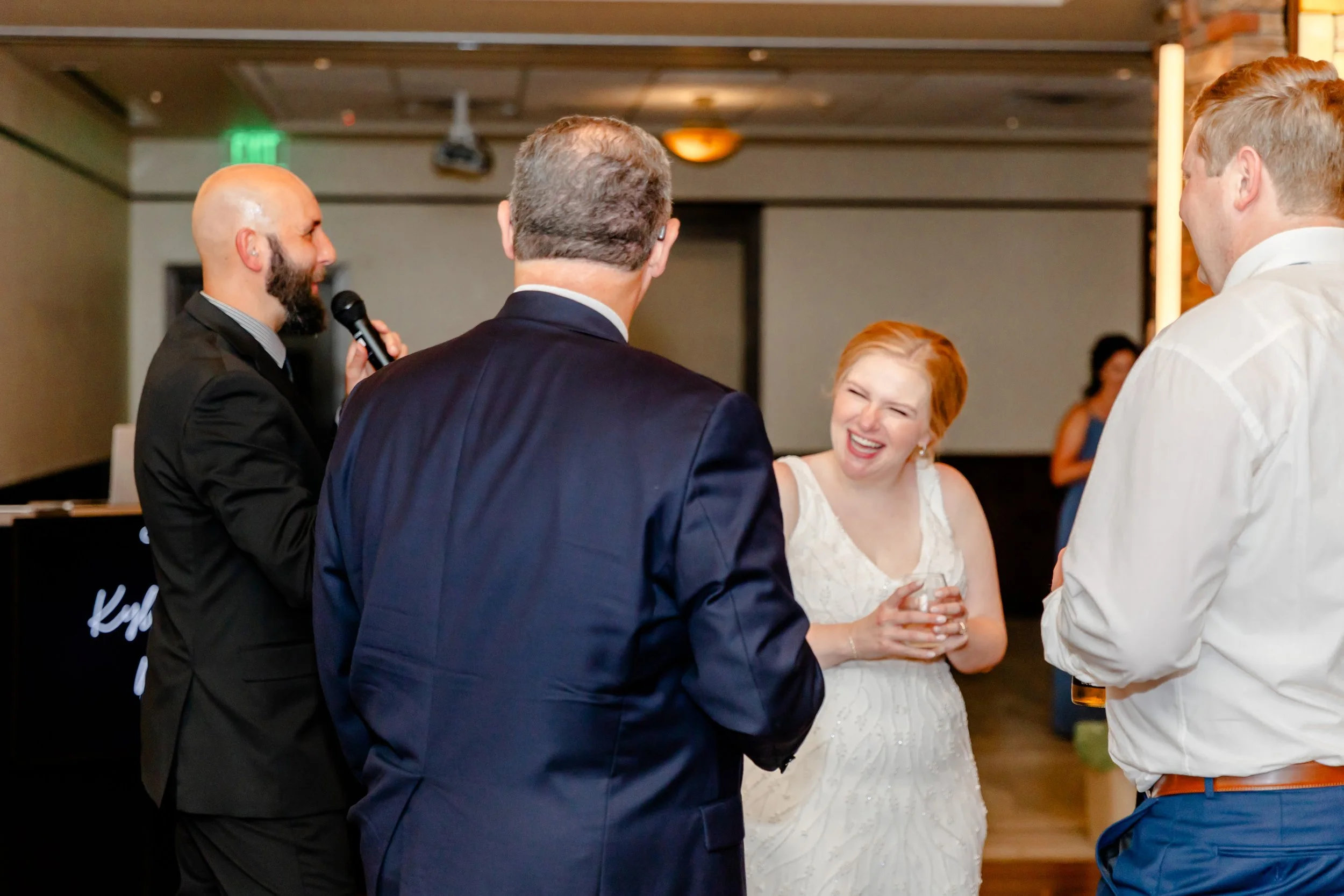 A group of five people, four men and one woman, are at a social event. The woman is wearing a white dress and smiling, holding a glass, while the men are dressed in formal attire and engaged in conversation, with one man holding a microphone.