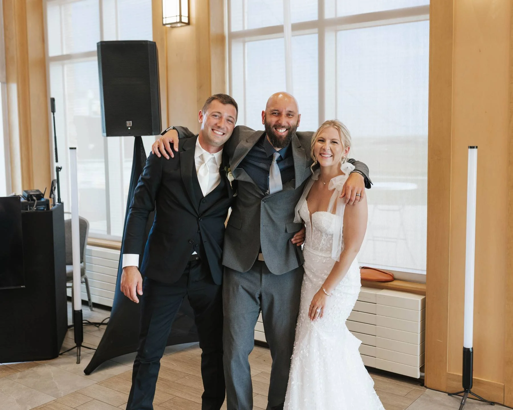 Three smiling people at a wedding: a groom in a black tuxedo, a bride in a white lace wedding gown, and a man in a gray suit, standing together with their arms around each other indoors in front of large windows.