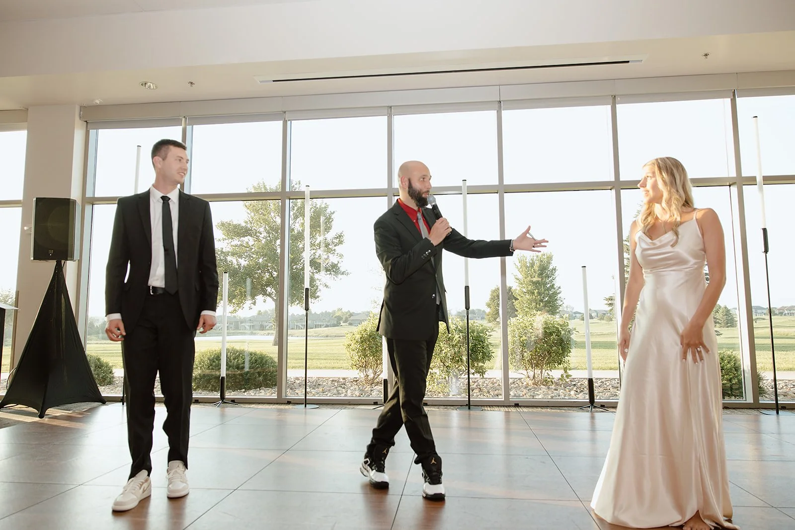 A wedding reception scene with a groom in a black suit, a woman in a cream-colored wedding dress, and a man in a black suit with a red shirt holding a microphone, standing on a dance floor with large windows showing trees and grass outside.