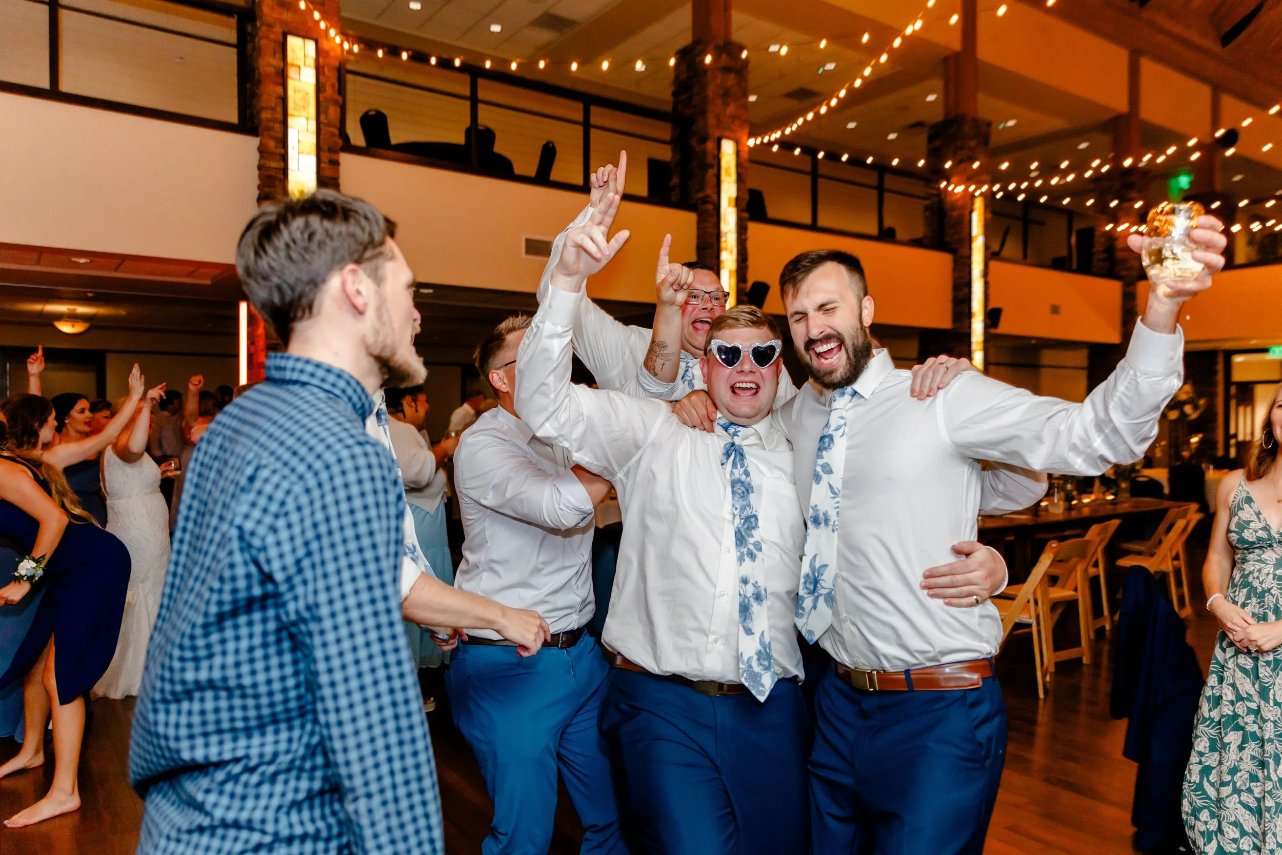Group of people celebrating and dancing at a party or wedding reception, some wearing sunglasses and informal attire, with colorful lighting and decorations in the background.