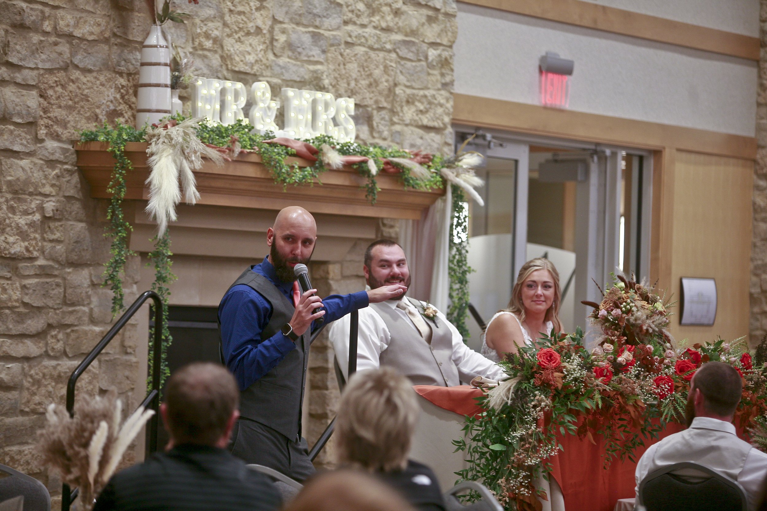 A man giving a speech at a wedding reception with a microphone, standing next to the bride and groom seated at a decorated table. The table is adorned with floral arrangements, and a sign above reads 'TR & TRS' on a decorated mantel. Guests are seated at tables, and the venue has a stone wall and wooden accents.