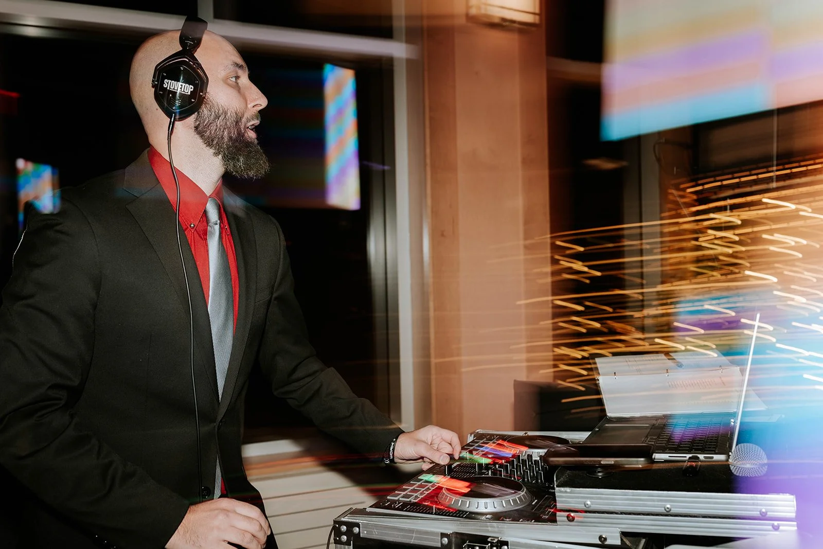 A DJ in a black suit, red shirt, and gray tie, wearing headphones, is operating DJ equipment with a laptop and a mixer in a dimly lit environment with colorful light streaks.