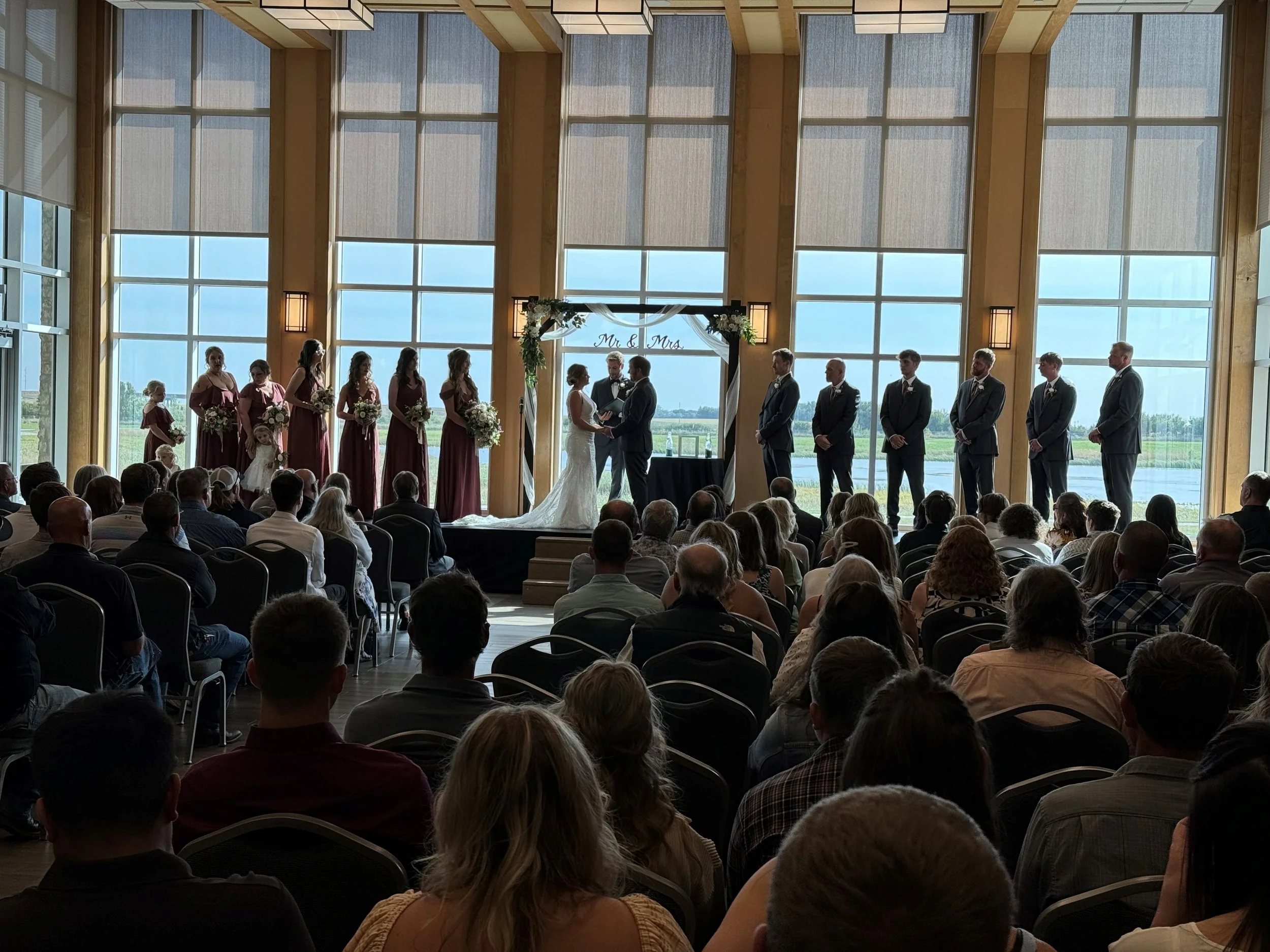 A wedding ceremony inside a large glass-fronted venue with a view of water and greenery outside. The bride and groom stand at the altar with officiant, surrounded by bridesmaids and groomsmen. The bridal party is dressed in formal attire, with the bridesmaids in matching dresses and the groomsmen in suits.