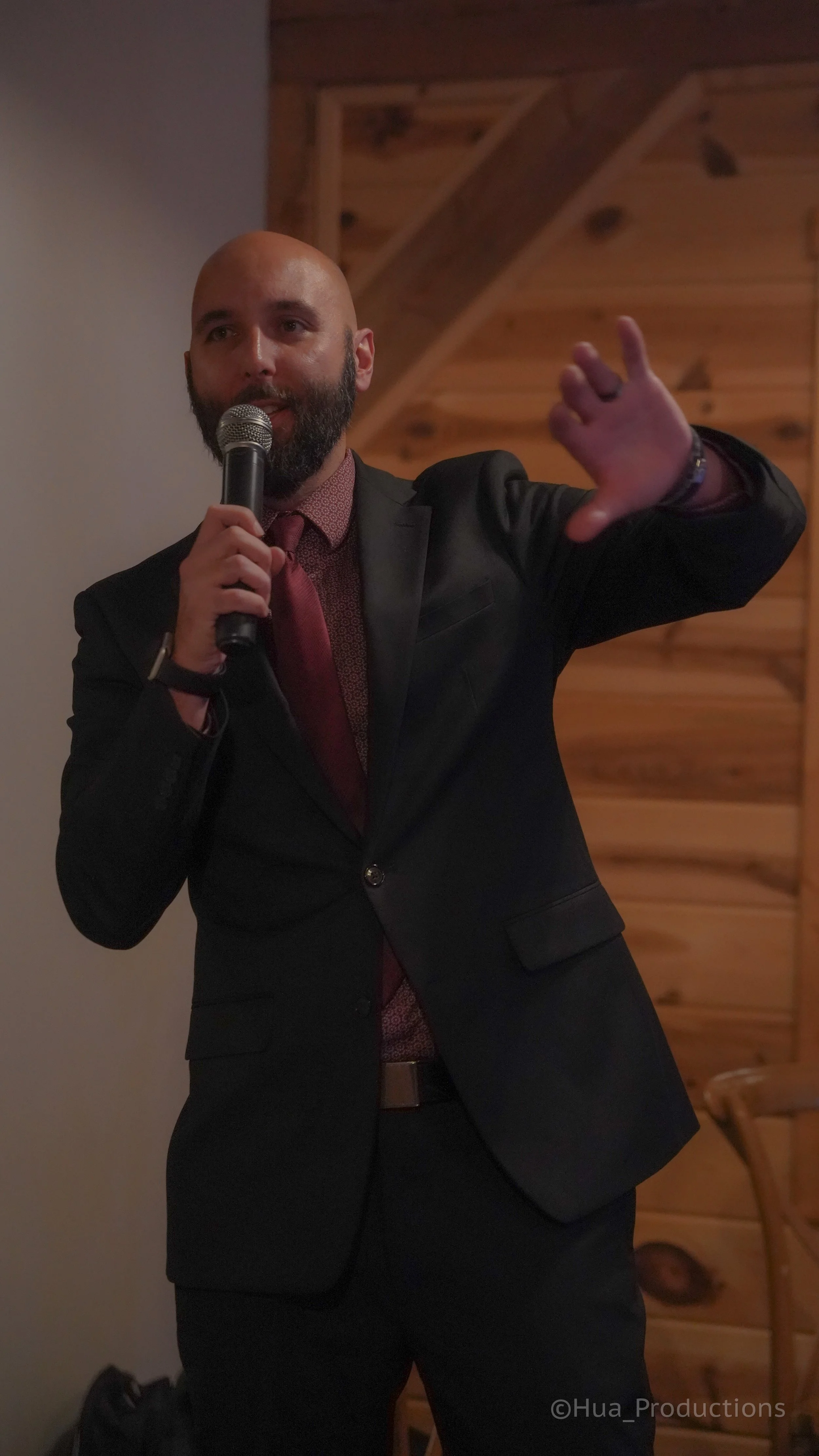 A man in a black suit and red tie holding a microphone, gesturing with his shoulder and arm extended, standing indoors with a wooden wall background.